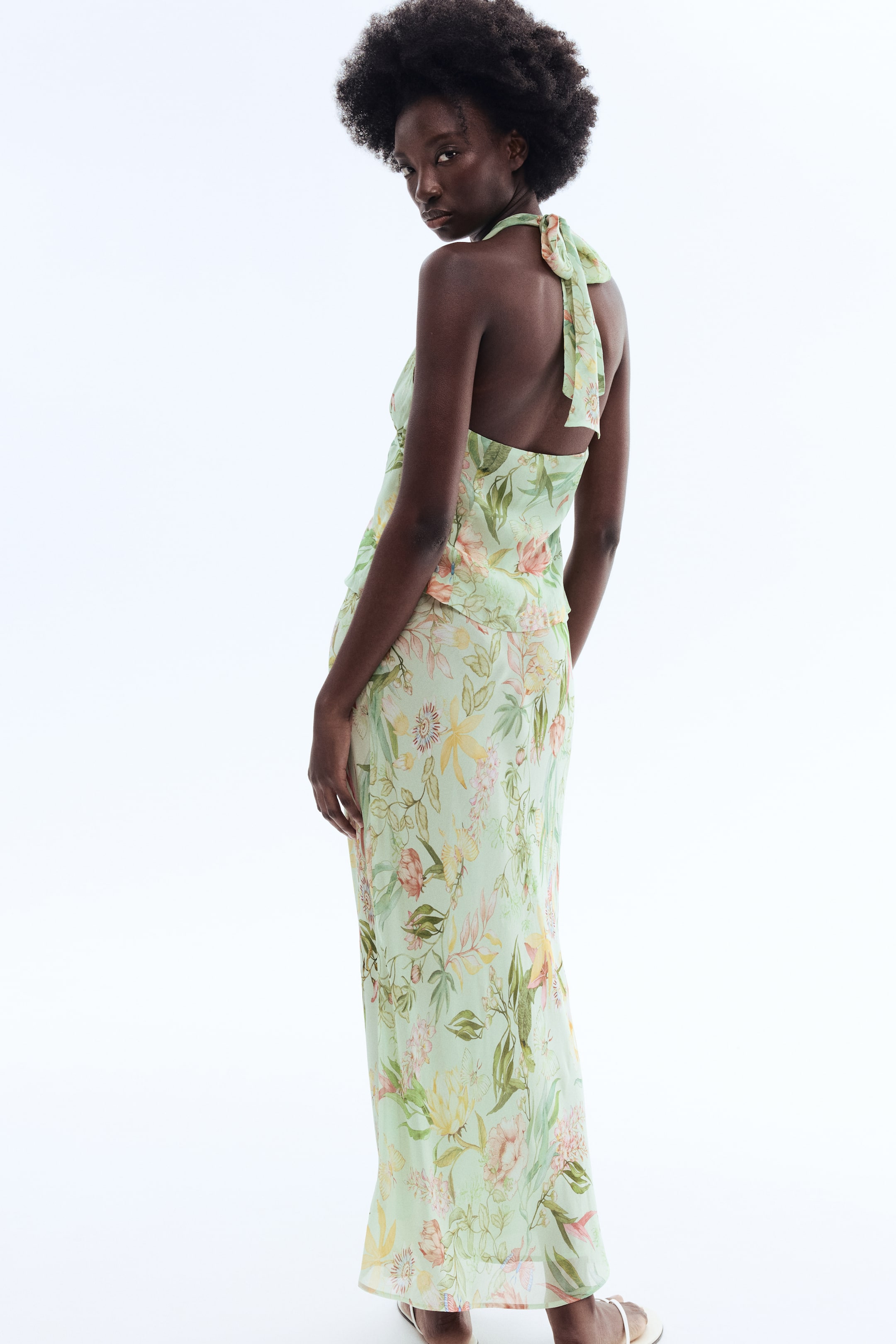 View larger image: A light green floral two-piece set is being shown from the back, featuring a halter top and a long skirt. The halter top's straps are forming a bow at the back of the neck, and both pieces are displaying a multi-coloured floral print with pink, yellow, and green elements.