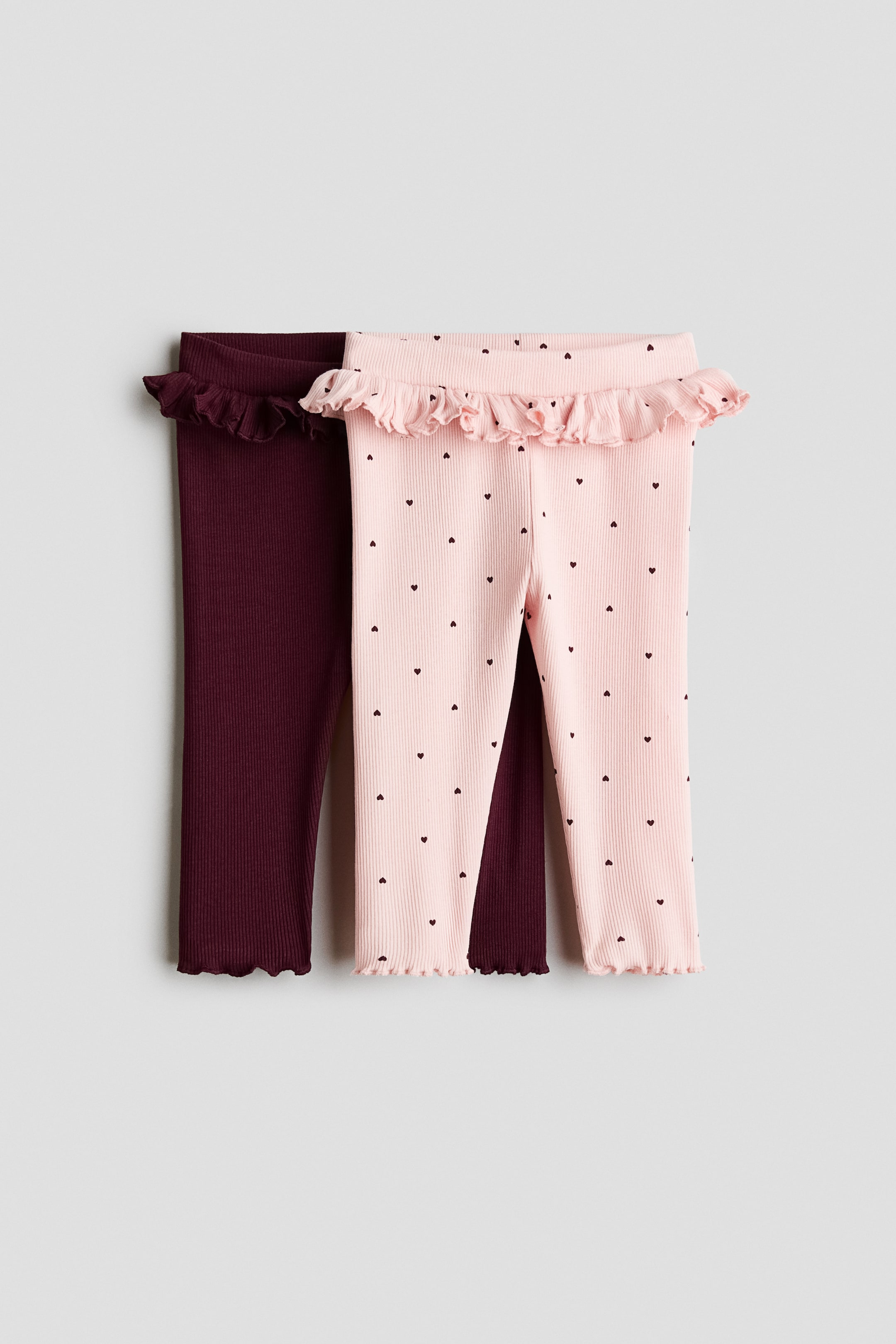 Größeres Bild ansehen: Zwei Paar gerippte Leggings sind übereinander getragen und jeweils von vorne zu sehen, mit gerüschten Bundabschlüssen und gerüschten Säumen. Das vorne liegende hellrosa Paar zeigt kleine dunkle Herzmuster, während das darüber liegende burgunderfarbene Paar ein unifarbenes Design aufweist.