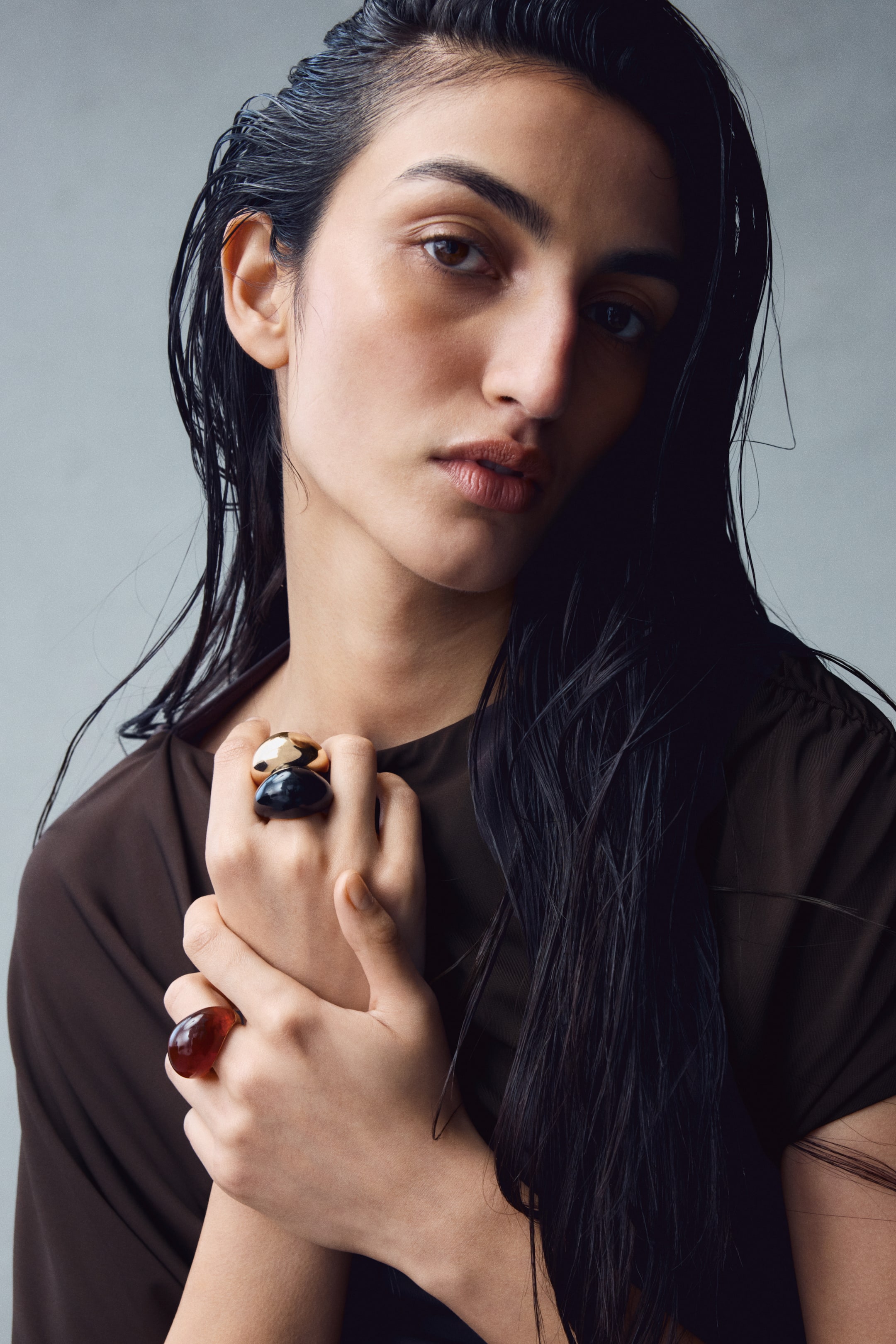 View larger image: Chunky rings in metallic gold, glossy black, and translucent amber finishes are worn on the model’s fingers, with their hands clasped together near the chest. The rings are styled with a dark brown, short-sleeved top.