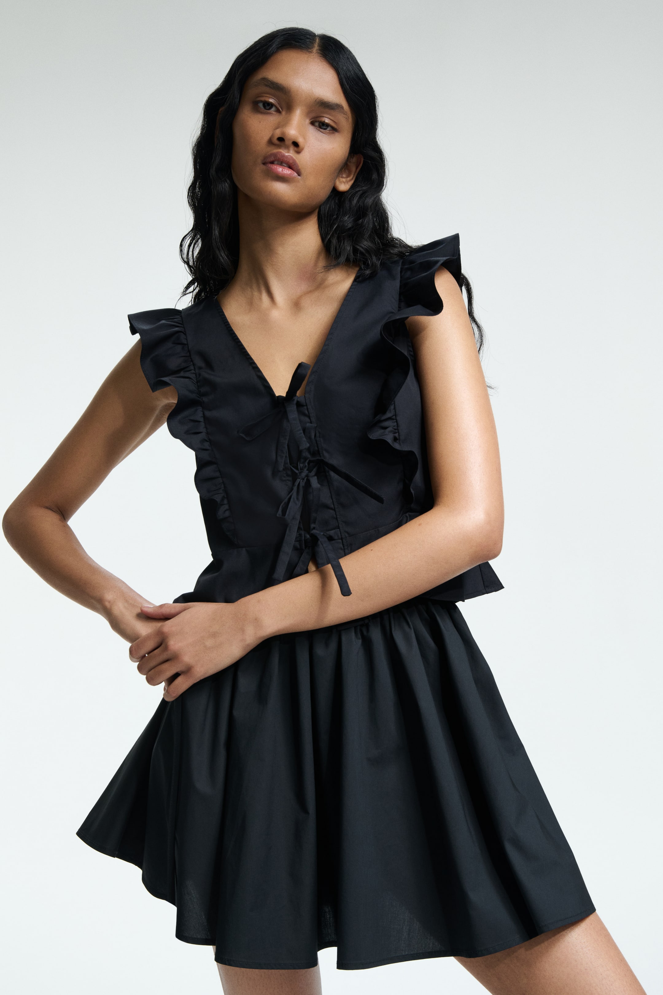 View larger image: A black two-piece outfit is being shown from the front, featuring a V-neck top with ruffled cap sleeves, vertical ruffle detailing on the front panels, and center lacing, paired with a gathered, flared mini skirt.