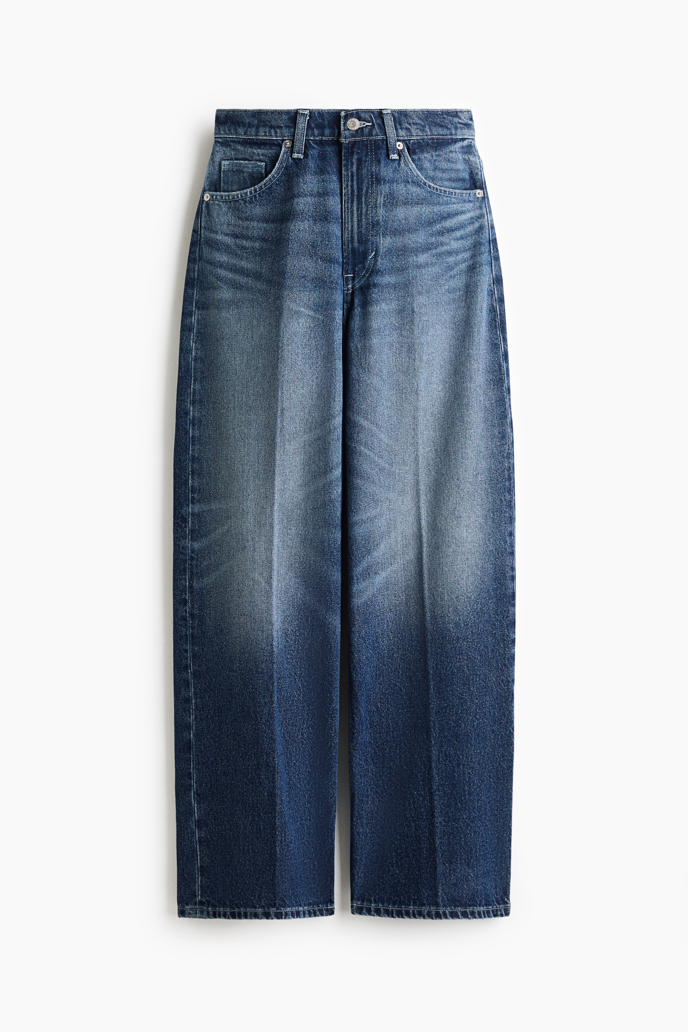 BlÃ¥ vide jeans med medium vask, frontslitasje og pressede folder ned langs beina bÃ¦res av modellen. Jeansene har en lÃ¸s, baggy passform og er stylet med en lagdelt hvit singlet og svarte sko med spiss tÃ¥.