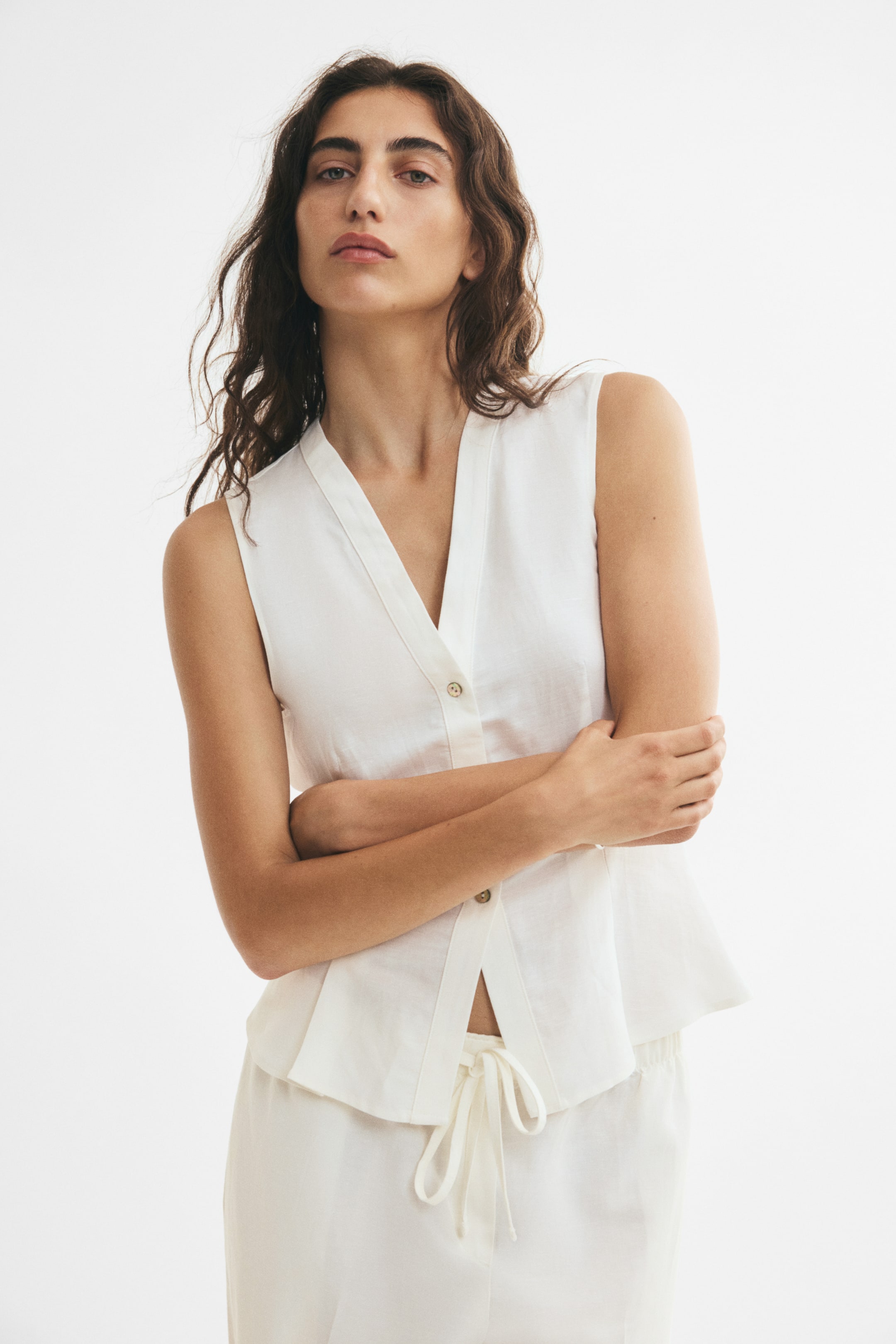 View larger image: A white, airy woven waistcoat made from a linen and viscose blend is worn by the model. It features a V-neckline, pearly buttons down to the waist, and shaping seams at the waist that open into a gently flared, pleated hem; the waistcoat is partially unbuttoned, revealing the drawstring of matching light-colored pants.