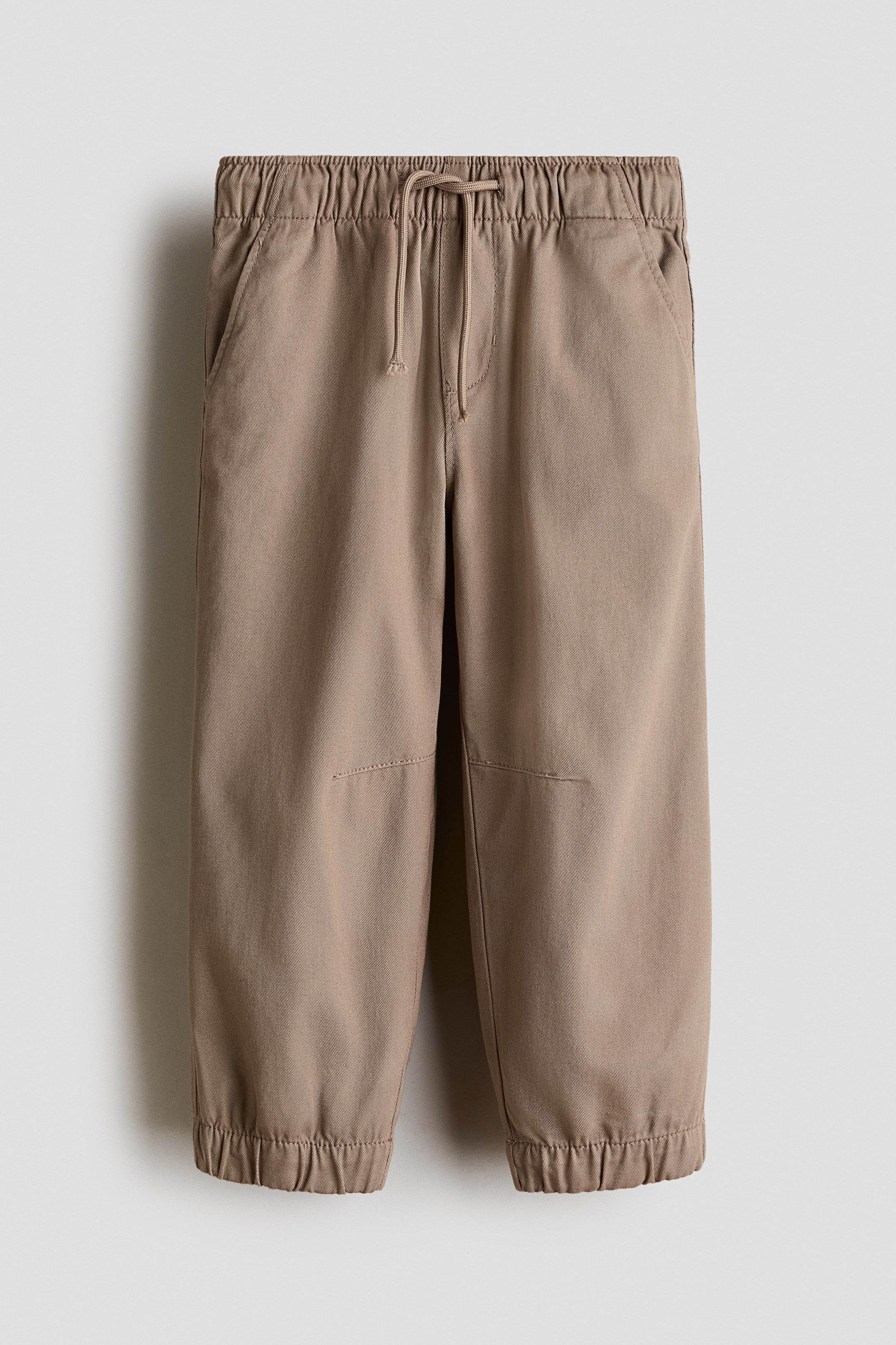 View larger image: Tan jogger pants feature an elasticated waistband with a drawstring closure, two slanted front pockets, stitched knee panels, and elasticated cuffs.