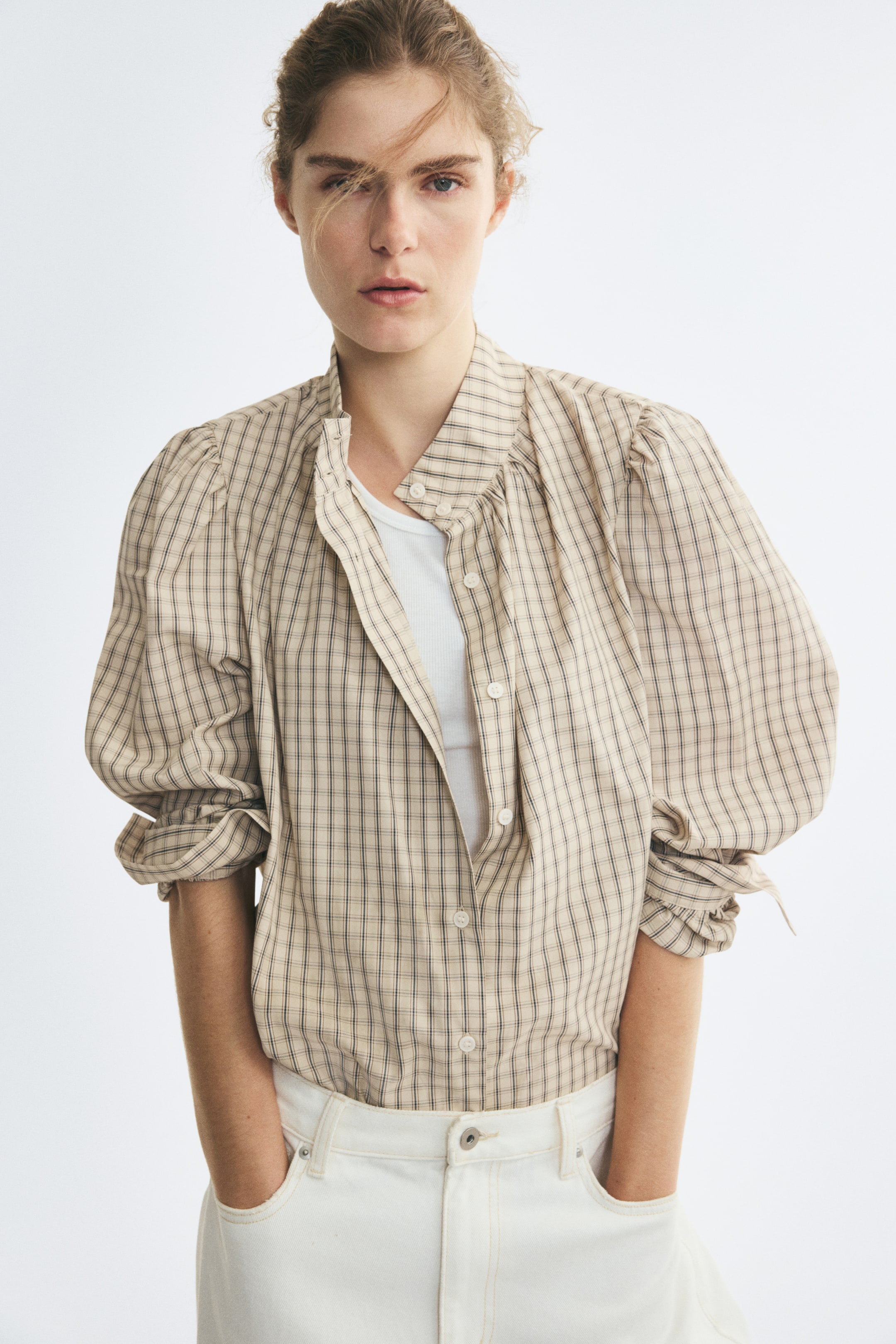 View larger image: The model is wearing a beige checked blouse, styled open over a white top and tucked into white trousers. The blouse features a stand collar, a button-front fastening, and voluminous puff sleeves pushed up on the forearms.