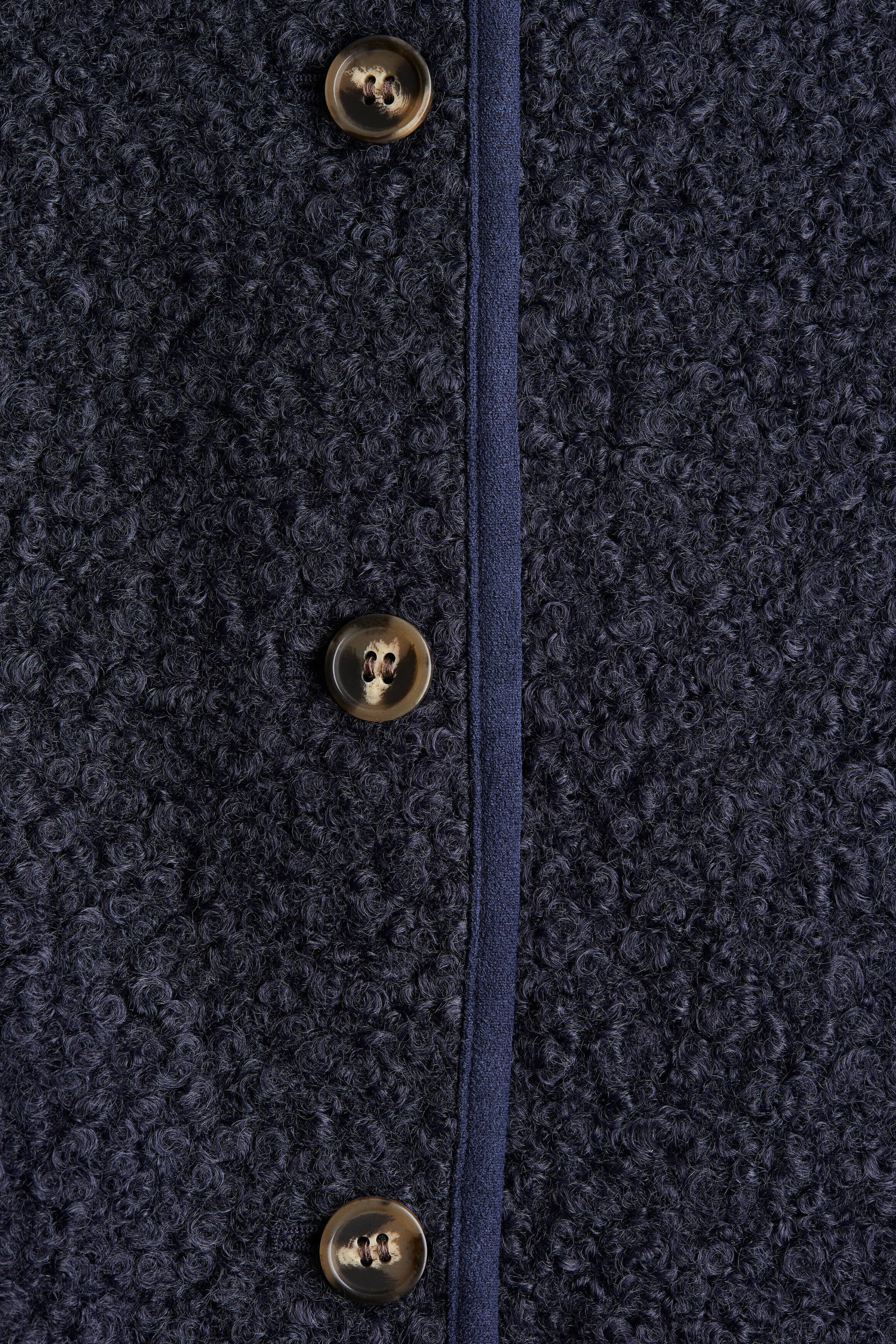 View larger image: The front placket of a dark blue short cardigan is visible, displaying its soft, curly teddy fabric. A smooth woven binding is running vertically down the center, and three round, mottled brown buttons are fastening the placket.
