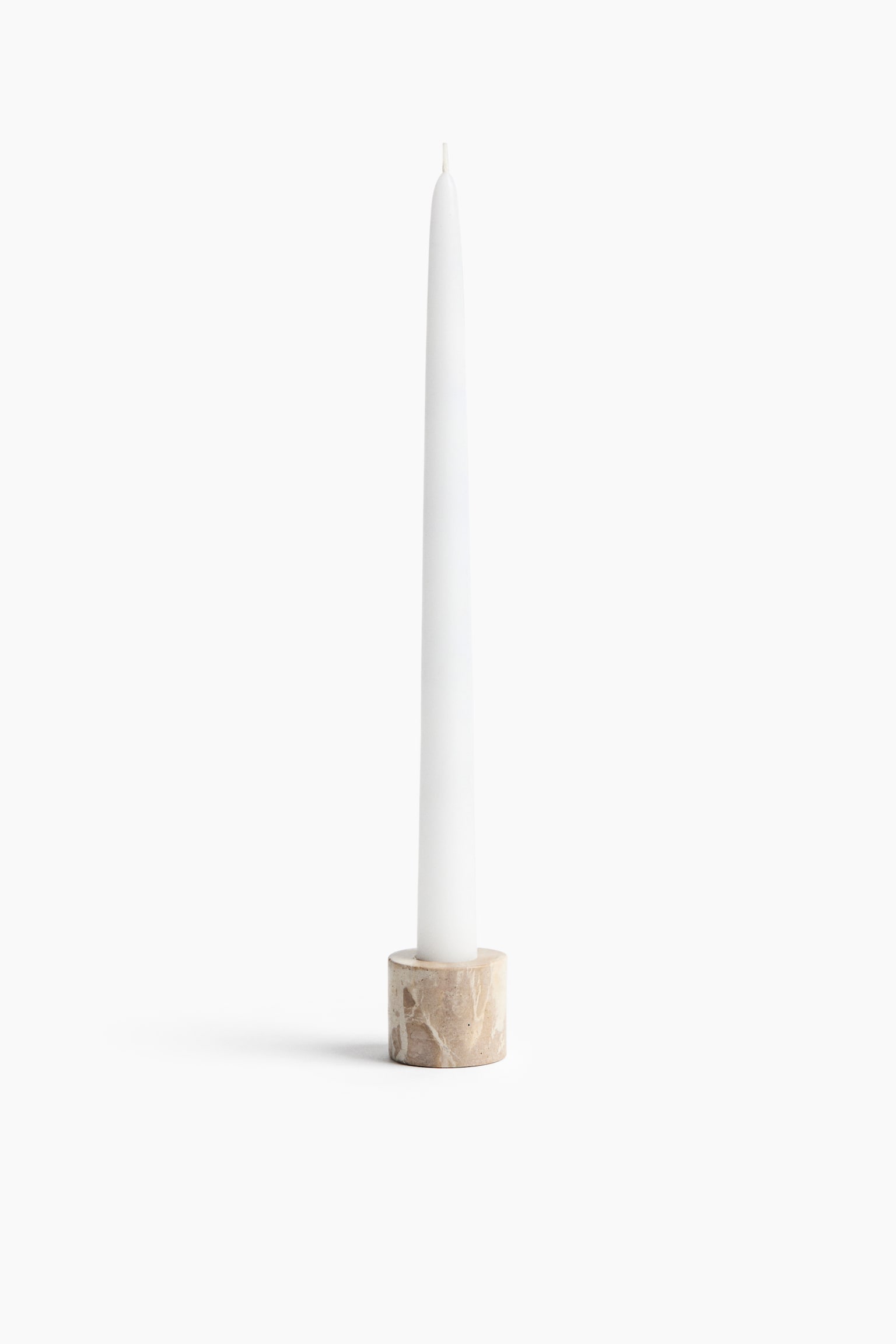 A small, cylindrical candle holder made of beige marbled stone with natural veining is holding a tall, white taper candle. The candle is standing vertically inside the holder, showing its unlit wick at the top.