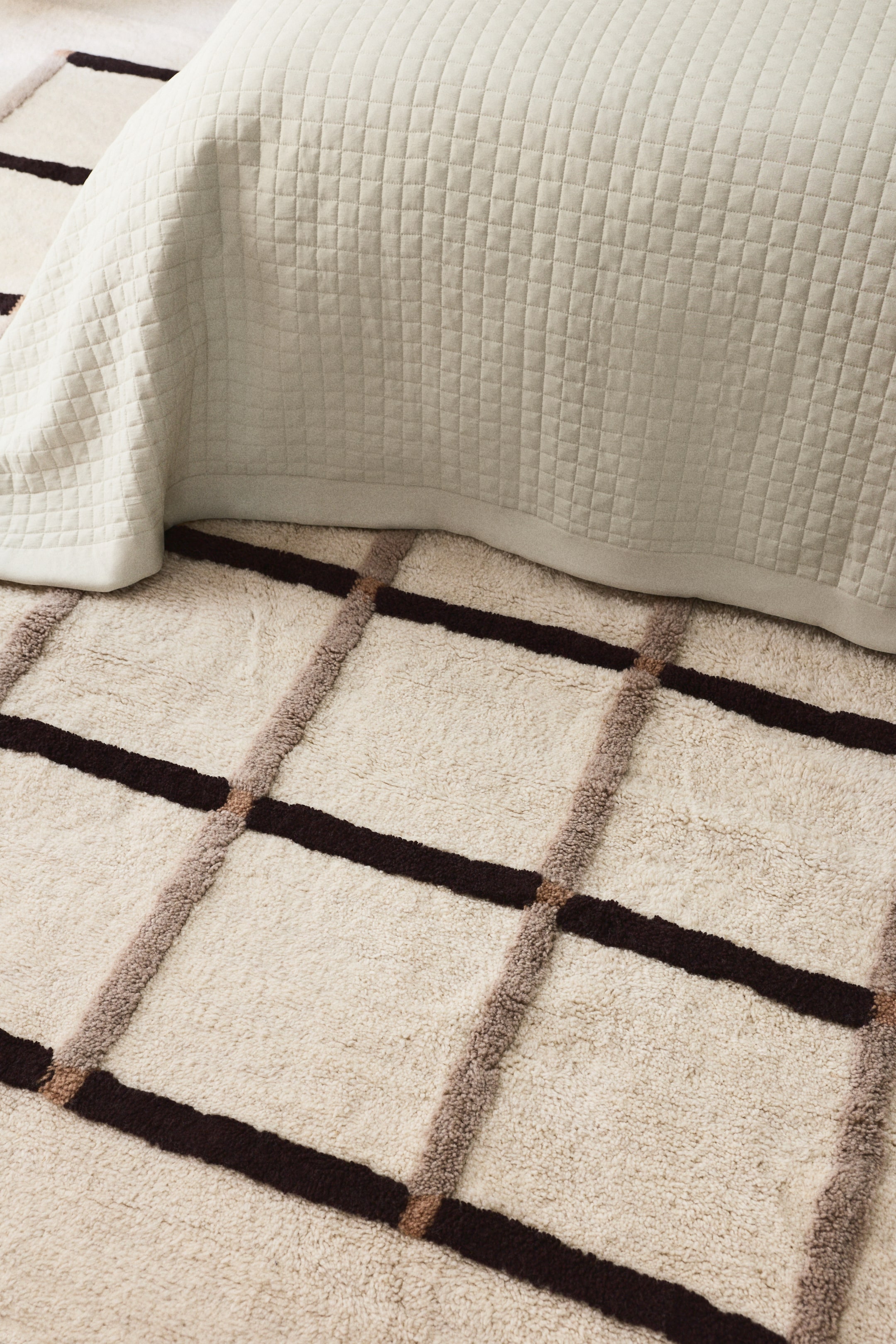 Nagyobb kép megtekintése: Egy krémszínű szőnyeg szabálytalan rácsmintát mutat vastag, texturált sötétbarna és világosbarna vonalakkal. Egy világos bézs steppelt takaró részben a szőnyegen fekszik, részben eltakarva annak mintáját.