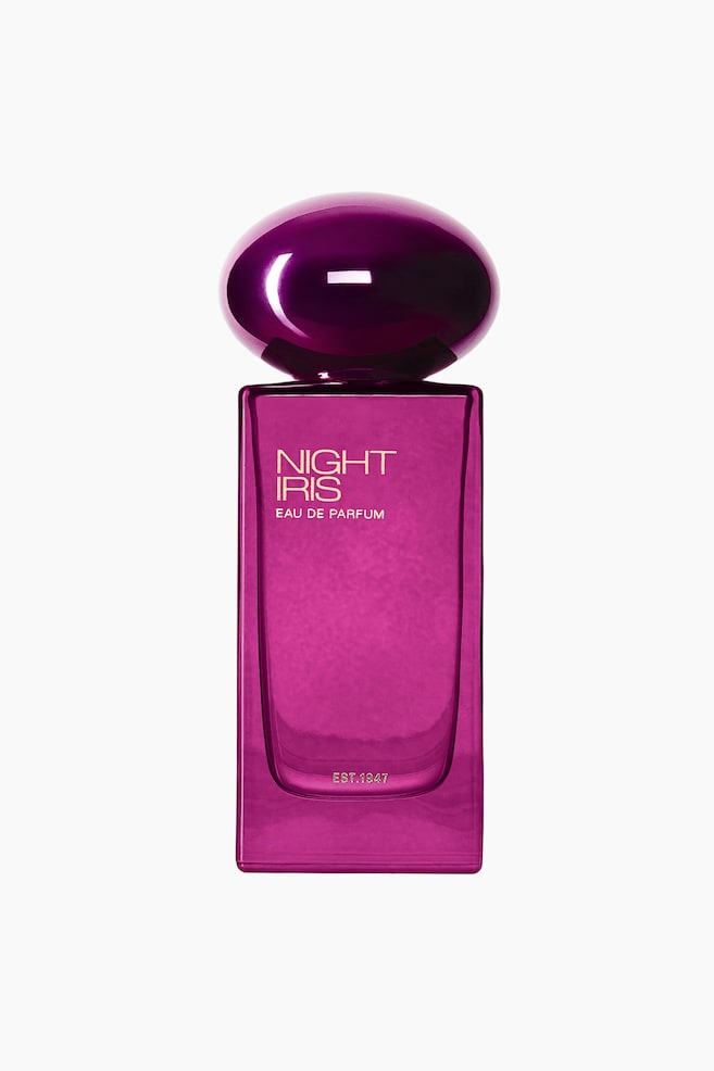 A bottle of Night Iris Eau de Parfum is shown from the front, featuring a rectangular, translucent magenta glass body topped with a glossy, dome-shaped metallic purple cap. The front of the bottle is printed with the text "NIGHT IRIS" and "EAU DE PARFUM" in gold lettering, with "EST. 1947" visible near the base. - 1