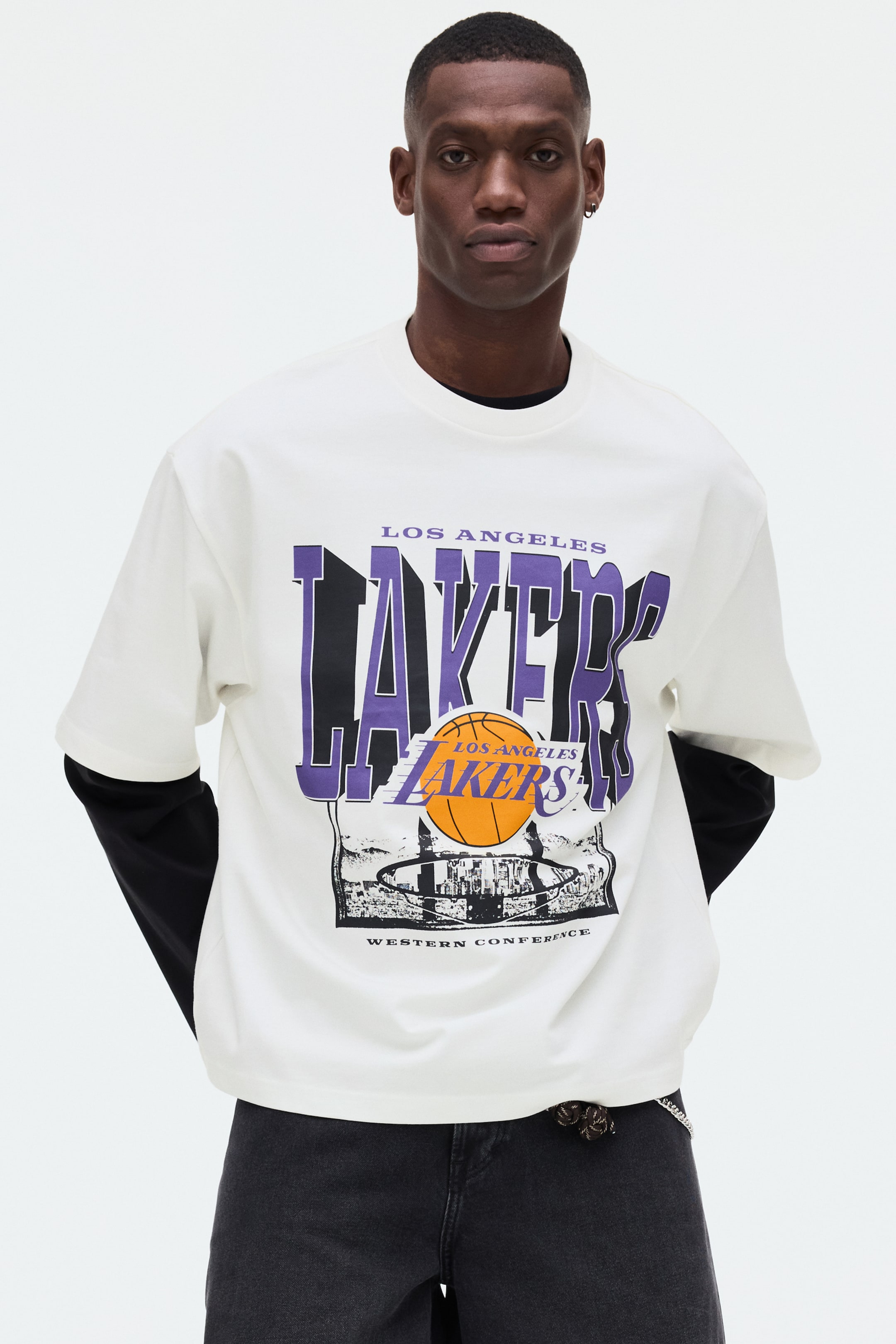 Visa större bild: En vit kortärmad t-shirt visar en stor grafik framtill med "LOS ANGELES" i lila, stiliserad "LAKERS"-text i lila och svart, en orange basketboll med texten "LOS ANGELES LAKERS", samt en svartvit stadionillustration med texten "WESTERN CONFERENCE". T-shirten bärs över en svart långärmad tröja, med de svarta ärmarna synliga undertill.