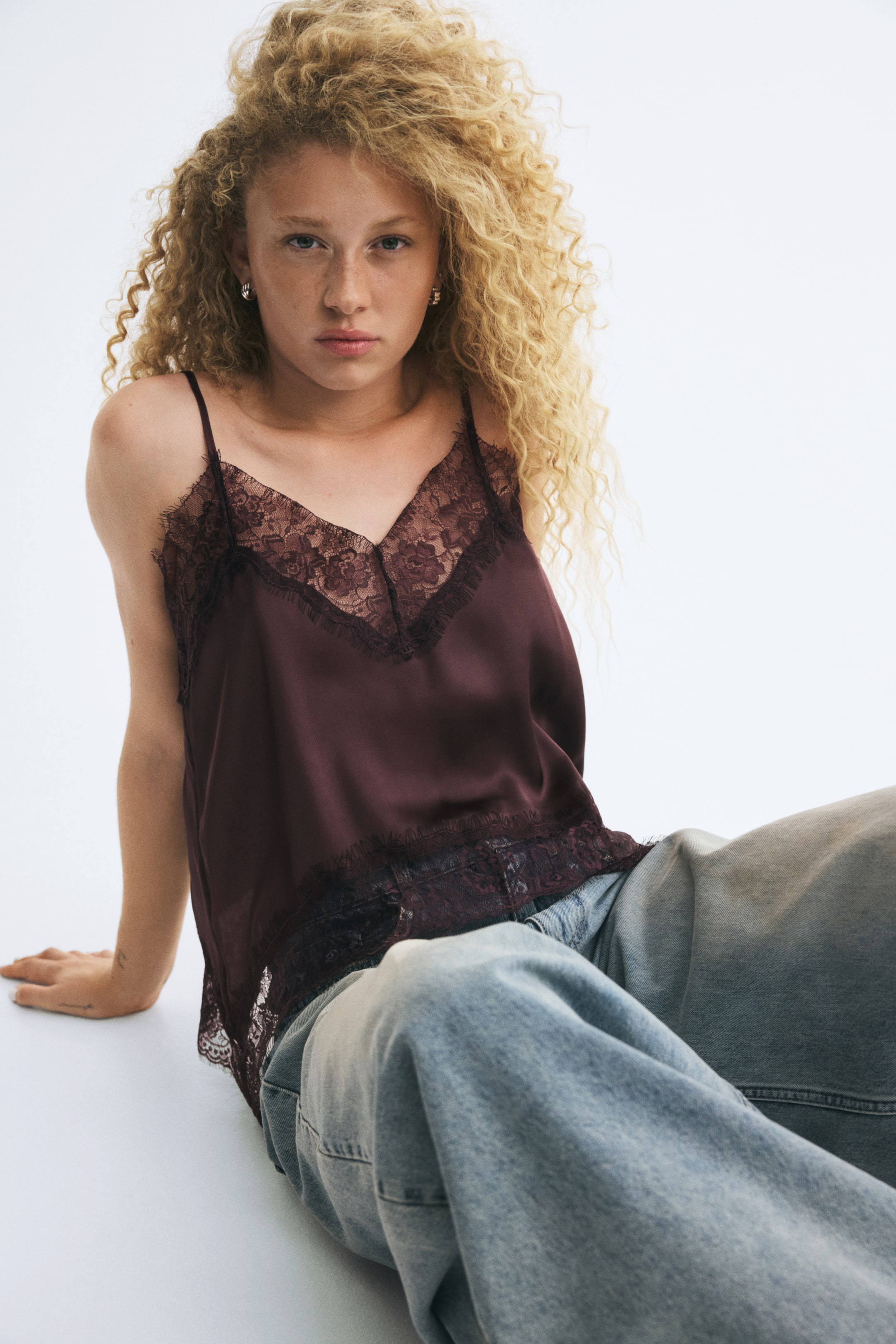 View larger image: A dark burgundy satin camisole featuring thin shoulder straps, a V-neckline, and delicate lace trim along the neck and hem is being worn by the model. The model is sitting while pairing the camisole with loose-fitting, light blue jeans.