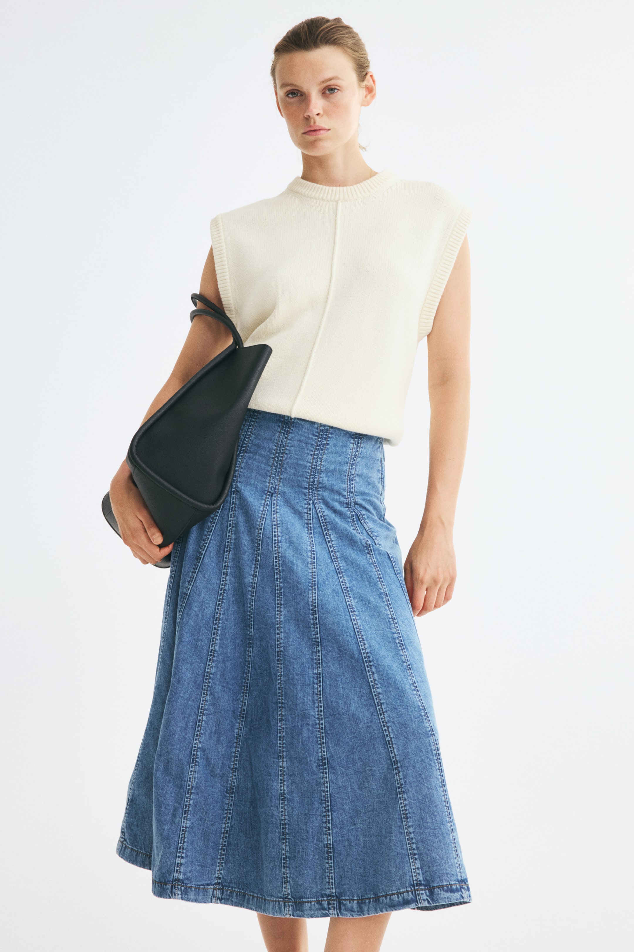 Voir une image plus grande: Une jupe en denim bleue longueur mi-mollet est portée par le mannequin, avec une taille ajustée et des empiècements à coutures verticales qui s’évasent en plis godets. La jupe est portée avec un haut en maille crème sans manches rentré à l’intérieur, et un sac à main noir est glissé sous le bras du mannequin.