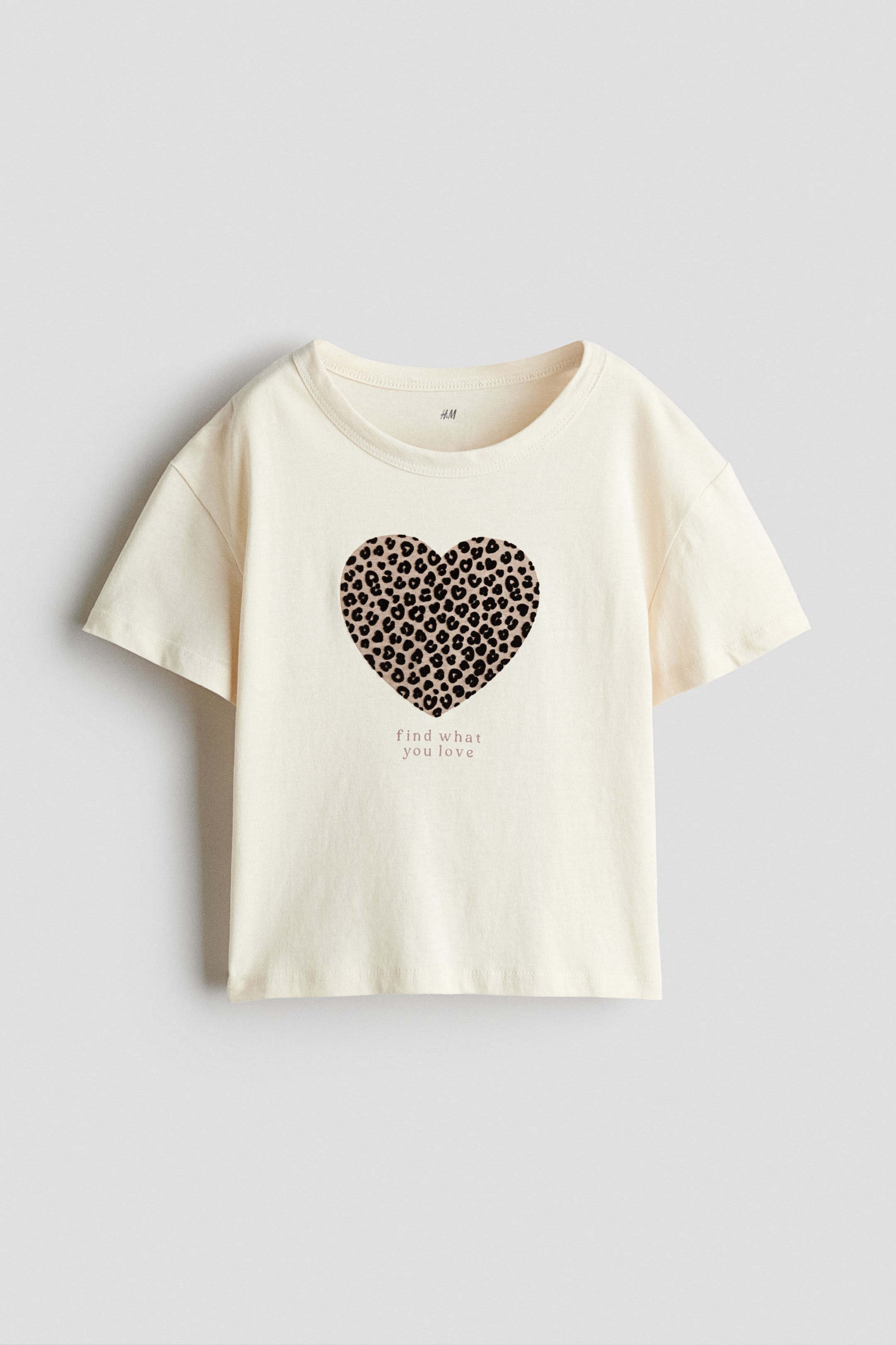 View larger image: A cream-colored cotton jersey tee is facing front, featuring a round neckline, dropped shoulders, and short sleeves. The tee displays a printed heart design with a leopard-print pattern above the lowercase text "find what you love" at the center.