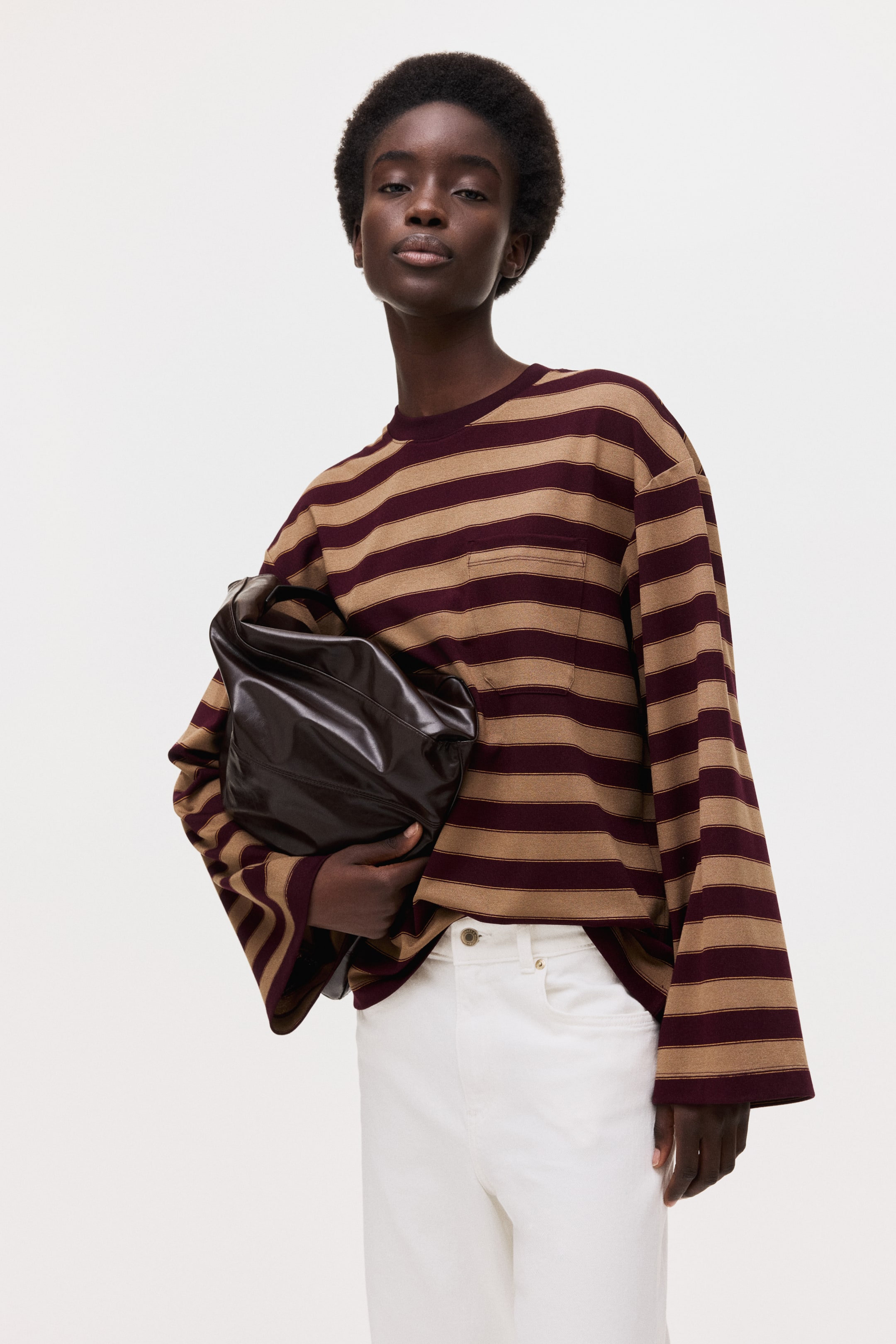 View larger image: A long-sleeved striped top features alternating horizontal stripes in burgundy and tan, presenting a relaxed, oversized fit with wide, flared sleeves, a crew neck and a single chest patch pocket. The top is styled over white trousers, with a dark brown gathered clutch held against the front of the top.