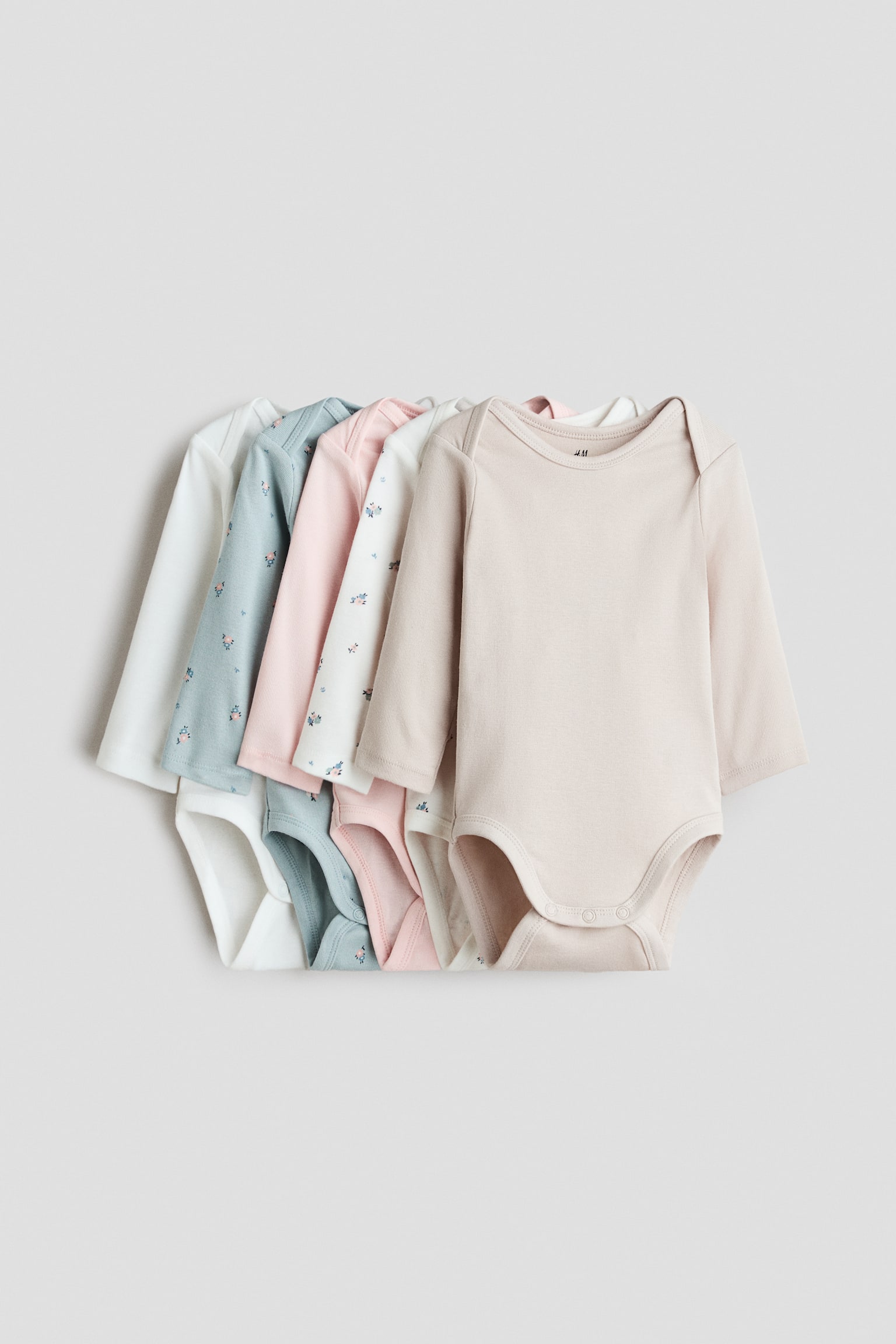 Five long-sleeve baby bodysuits are laid flat in a layered, overlapping stack, facing forward to showcase their envelope necklines and press-stud fastenings at the bottom. The assortment includes solid light beige, solid light pink and solid white styles, alongside two floral-printed designs in off-white and dusty blue.