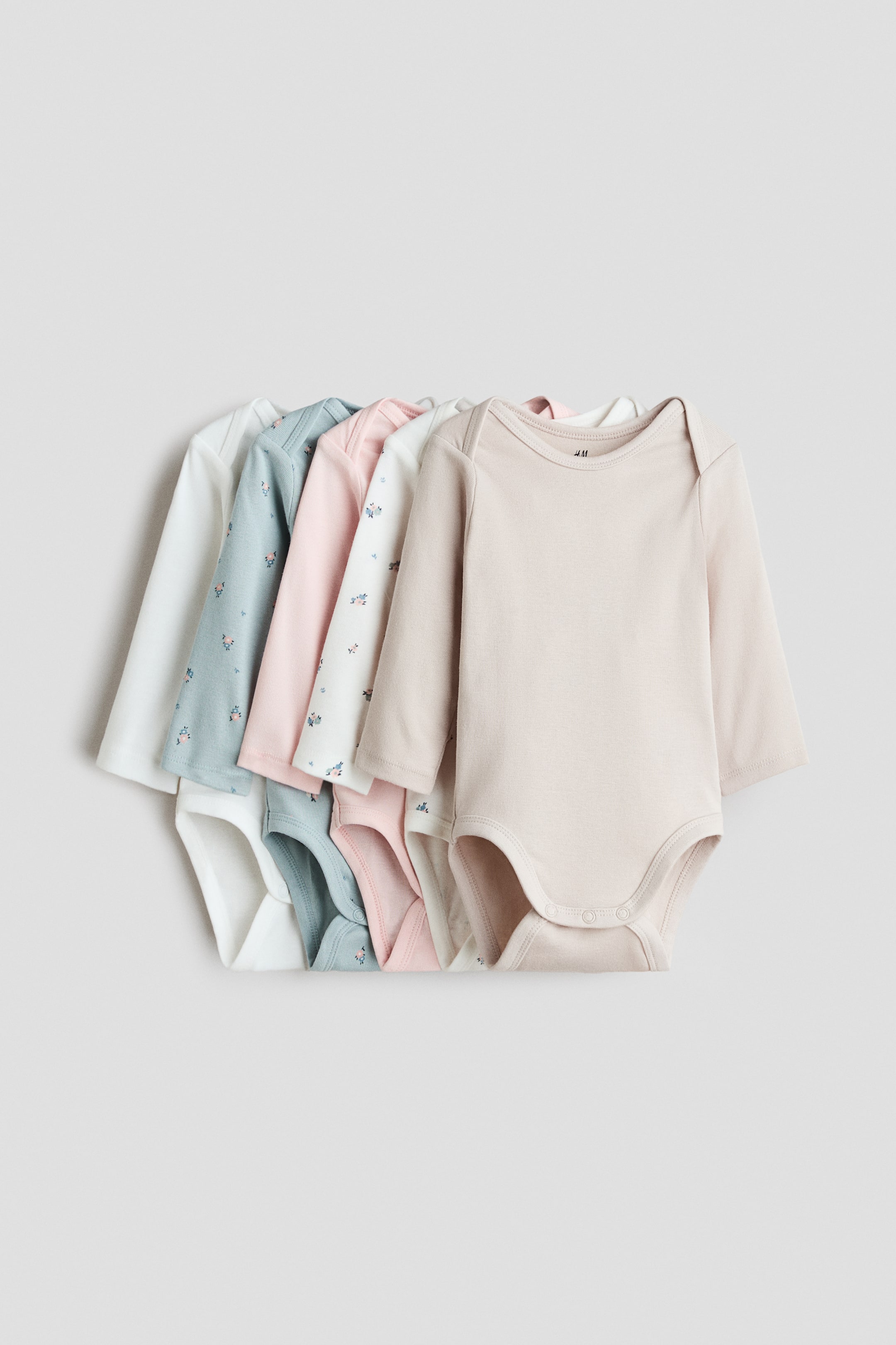 View larger image: Five long-sleeve baby bodysuits are laid flat in a layered, overlapping stack facing forward, showcasing their envelope necklines and press-stud closures at the bottom. The assortment includes solid light beige, solid light pink, and solid white styles, alongside two floral-printed designs in off-white and dusty blue.