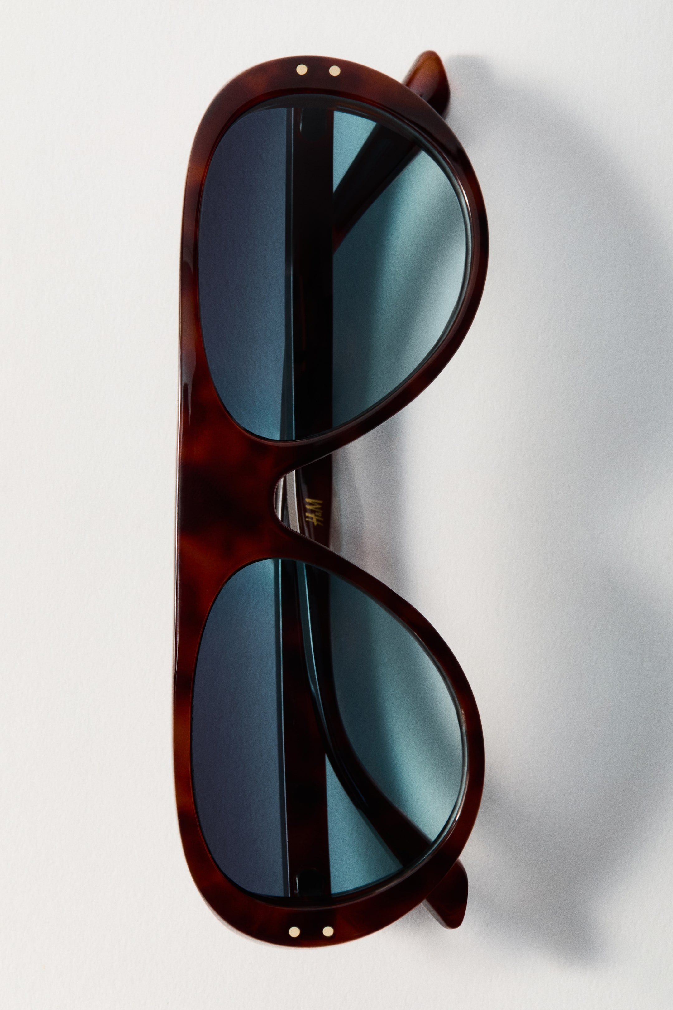 View larger image: Shield sunglasses feature a wide, tortoise shell plastic frame with small gold circular accents at the top outer corners and blue-tinted plastic lenses.