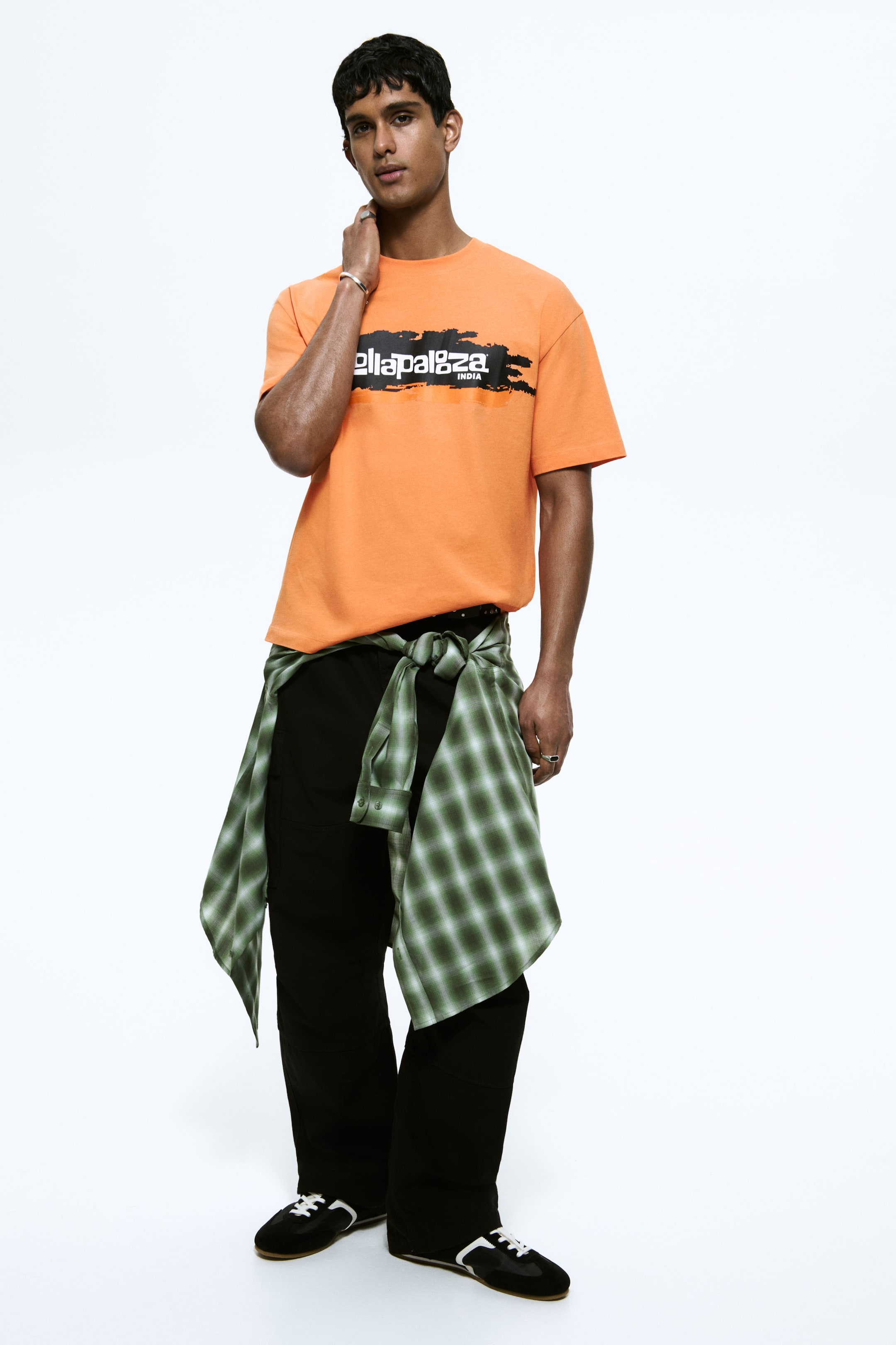 View larger image: The model is wearing an orange T-shirt with a black and orange graphic print. The T-shirt features a round, ribbed neckline, dropped shoulders, a straight hem, and a relaxed fit. A green and white checked shirt is tied around the model’s waist to style the T-shirt.