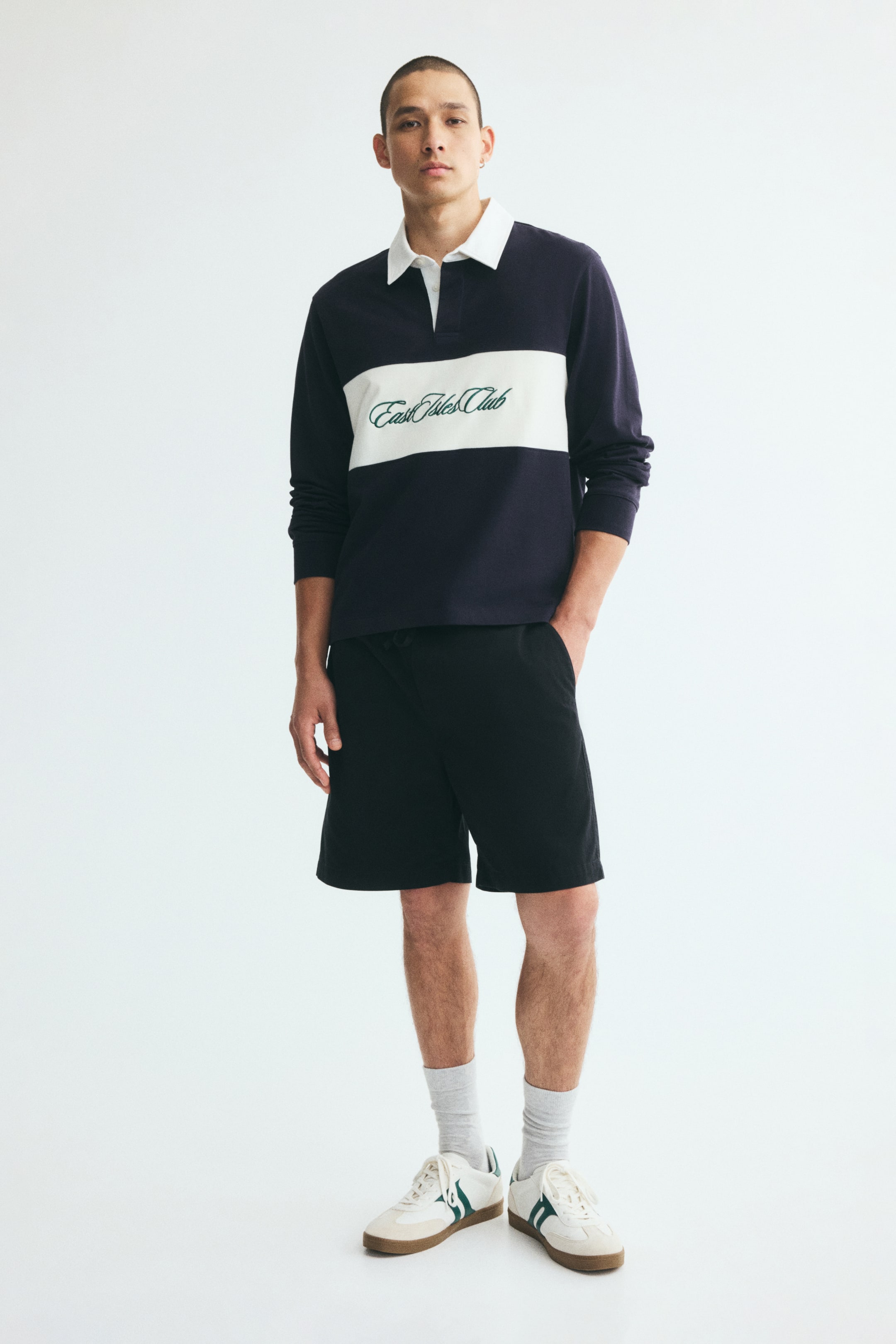 View larger image: Black cotton twill shorts with a relaxed fit are worn by the model, who is standing with one hand tucked into a diagonal side pocket. The shorts are styled with a navy and white long-sleeve polo shirt, gray crew socks, and white sneakers.
