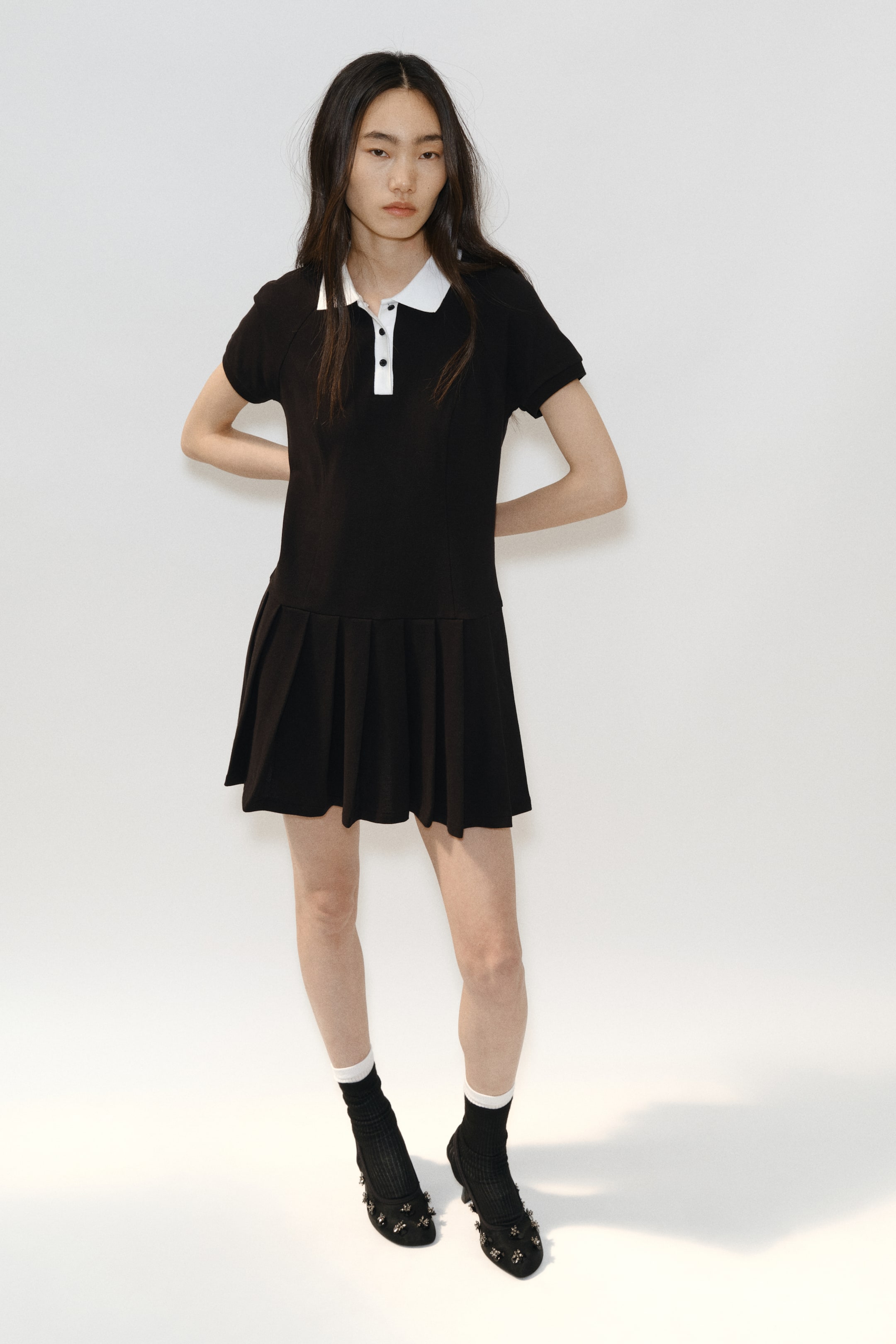 拡大画像を表示: 黒のポロドレスは、白い襟と前立てに黒のボタンが付いており、短い袖とプリーツスカートが正面を向いています。