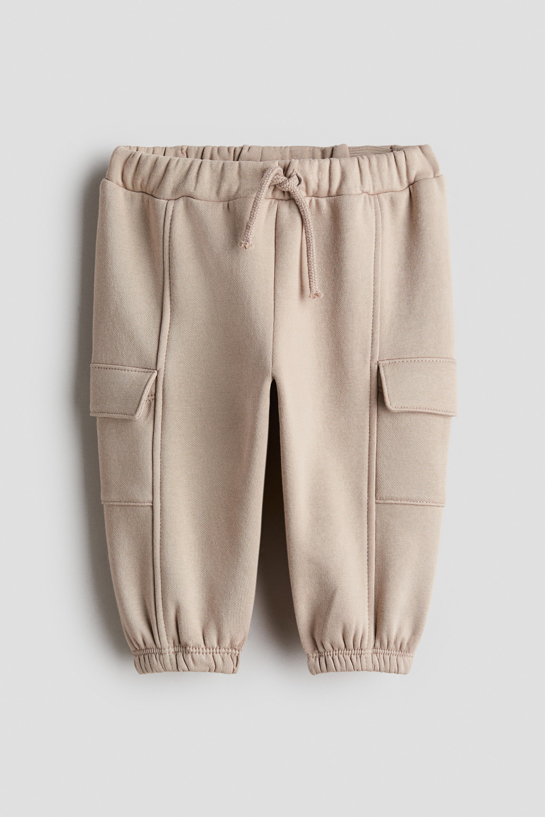 View larger image: A pair of beige cargo pants featuring an elastic drawstring waistband, prominent vertical seams running down the front of each leg, and two flap cargo pockets on the mid-thigh, with elastic cuffs finishing the ankles.