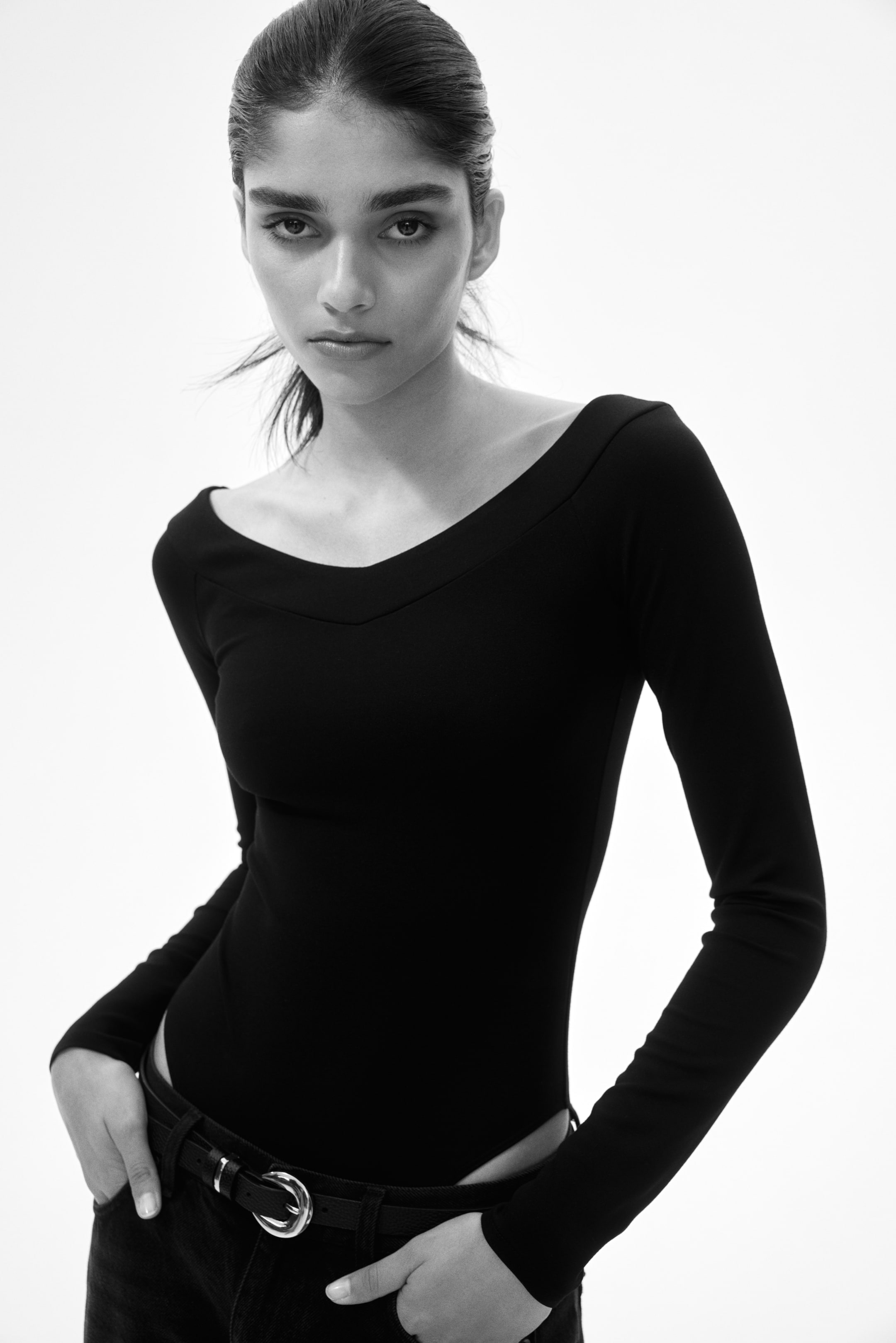 View larger image: This black long-sleeved bodysuit showcases a wide boat neckline accentuated by a V-shaped seam detail. Its sleek, form-fitting design is neatly tucked into dark trousers.