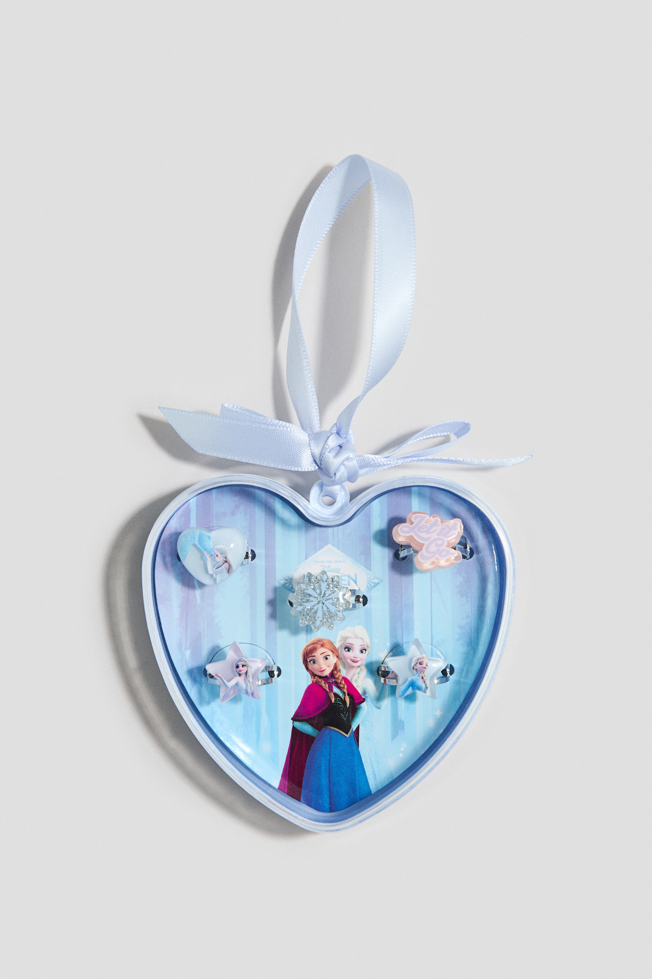 View larger image: A heart-shaped, light blue plastic box, tied at its top with a light blue satin ribbon, displays a set of metal and plastic rings. Several rings, showing a variety of designs including character images, snowflakes and stars, are arranged inside the box.