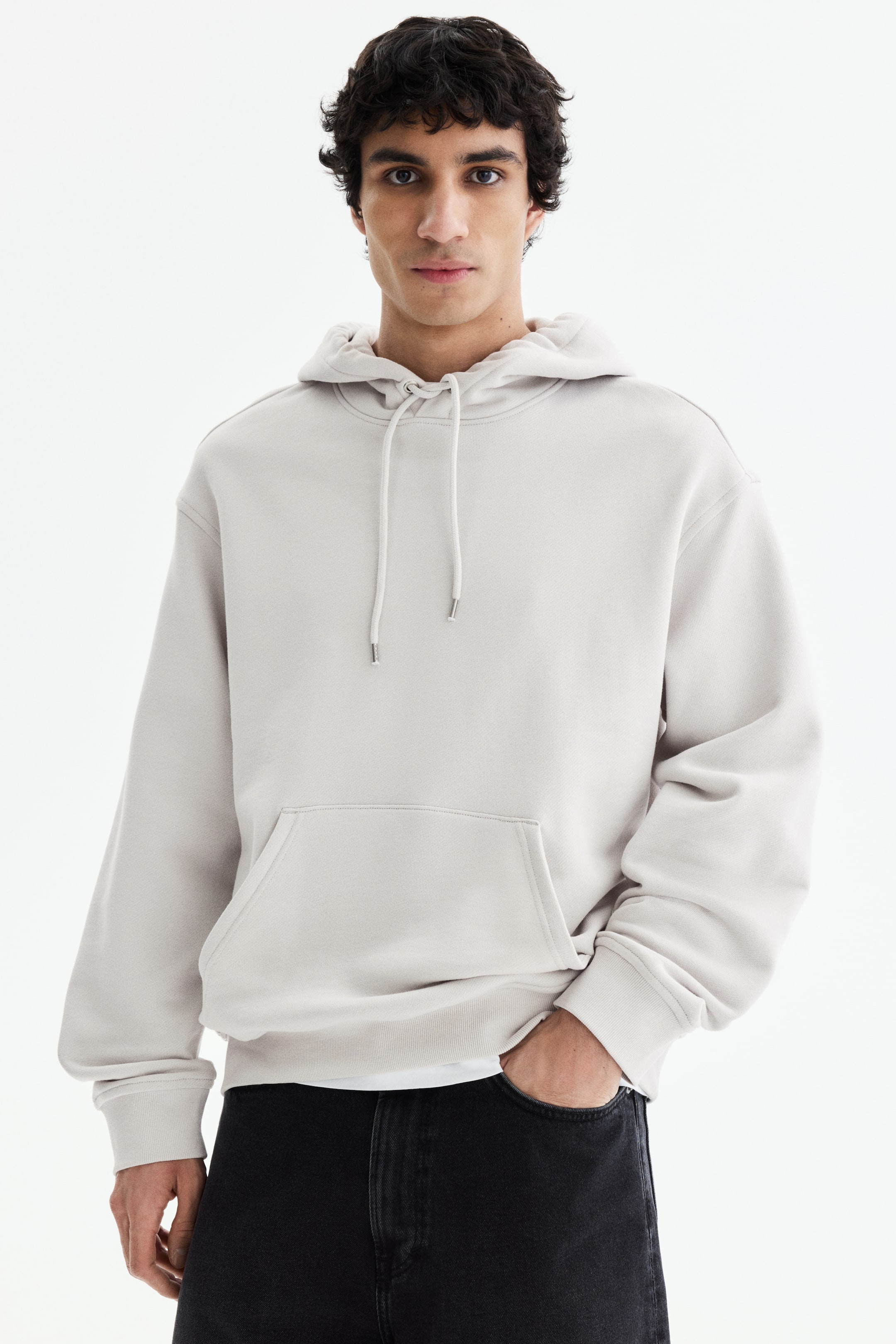 View larger image: A light beige hoodie is facing forward, featuring a hood with white drawstrings tipped with silver, a large kangaroo pocket, and long sleeves with ribbed cuffs. A white t-shirt is peeking out from under its ribbed hem.