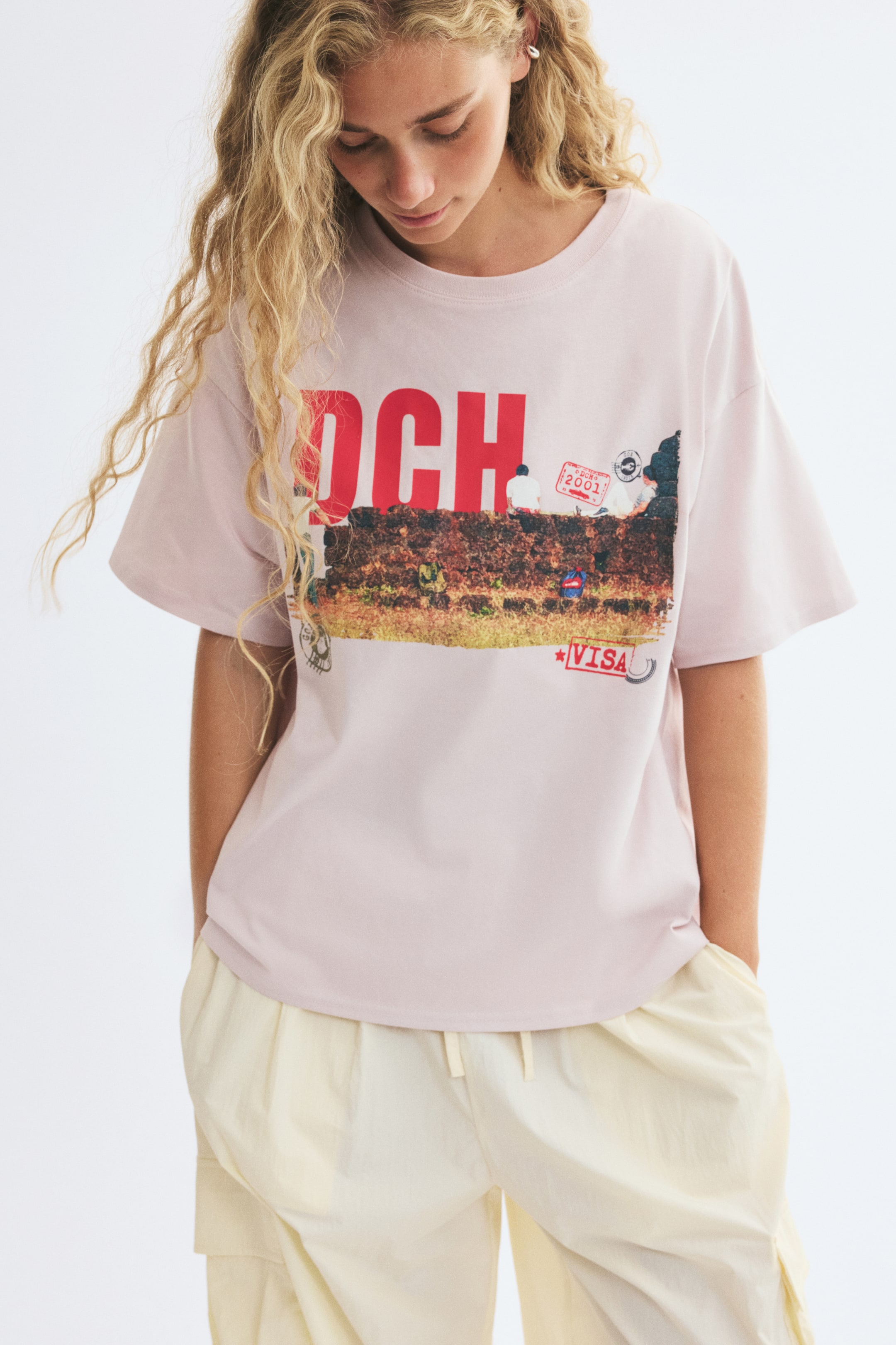 View larger image: An oversized light pink cotton T-shirt with a round neckline, dropped shoulders, and a colourful graphic print on the chest is worn by the model. The T-shirt is styled with light cream-coloured, loose-fitting trousers.