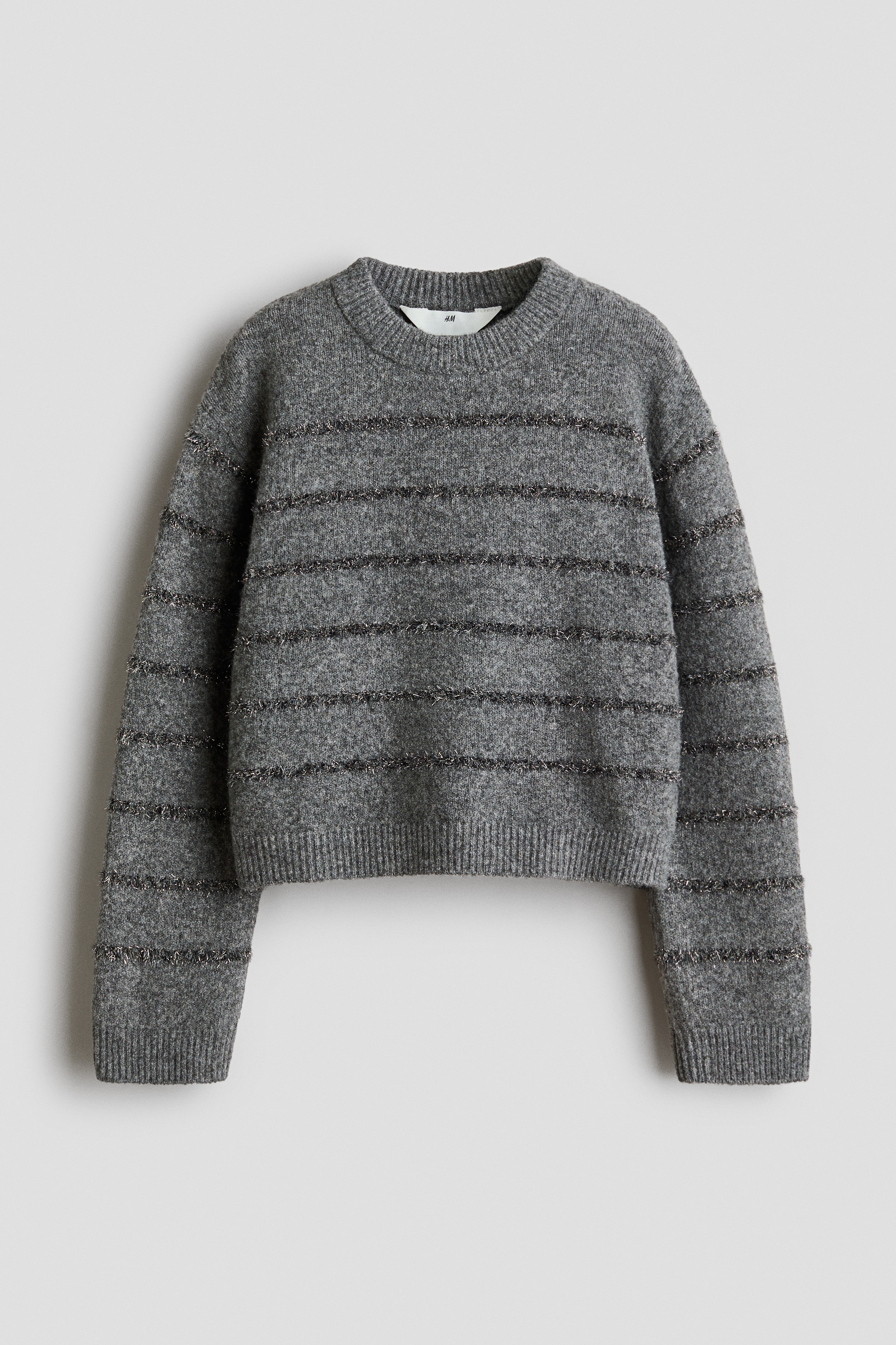 Un pull en maille gris est présenté de face, avec un col rond côtelé, des épaules légèrement tombantes, ainsi que des poignets et un ourlet côtelés. Le design est rehaussé de rayures horizontales en fils métalliques texturés et scintillants sur l’ensemble de la maille douce.