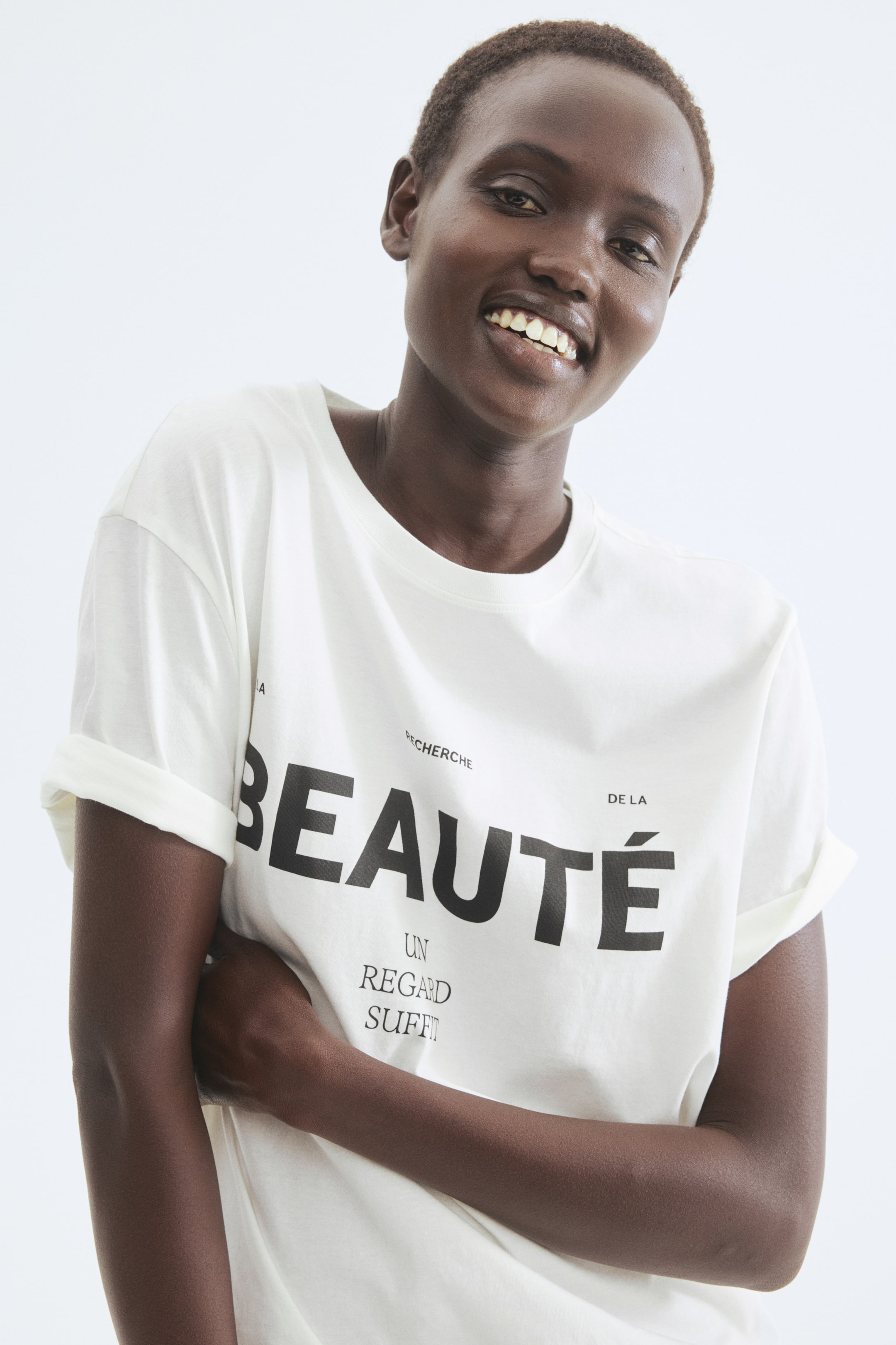 View larger image: A white t-shirt features a round neck and rolled short sleeves. The front displays a black text print that reads "RECHERCHE DE LA BEAUTÉ UN REGARD SUFFIT."