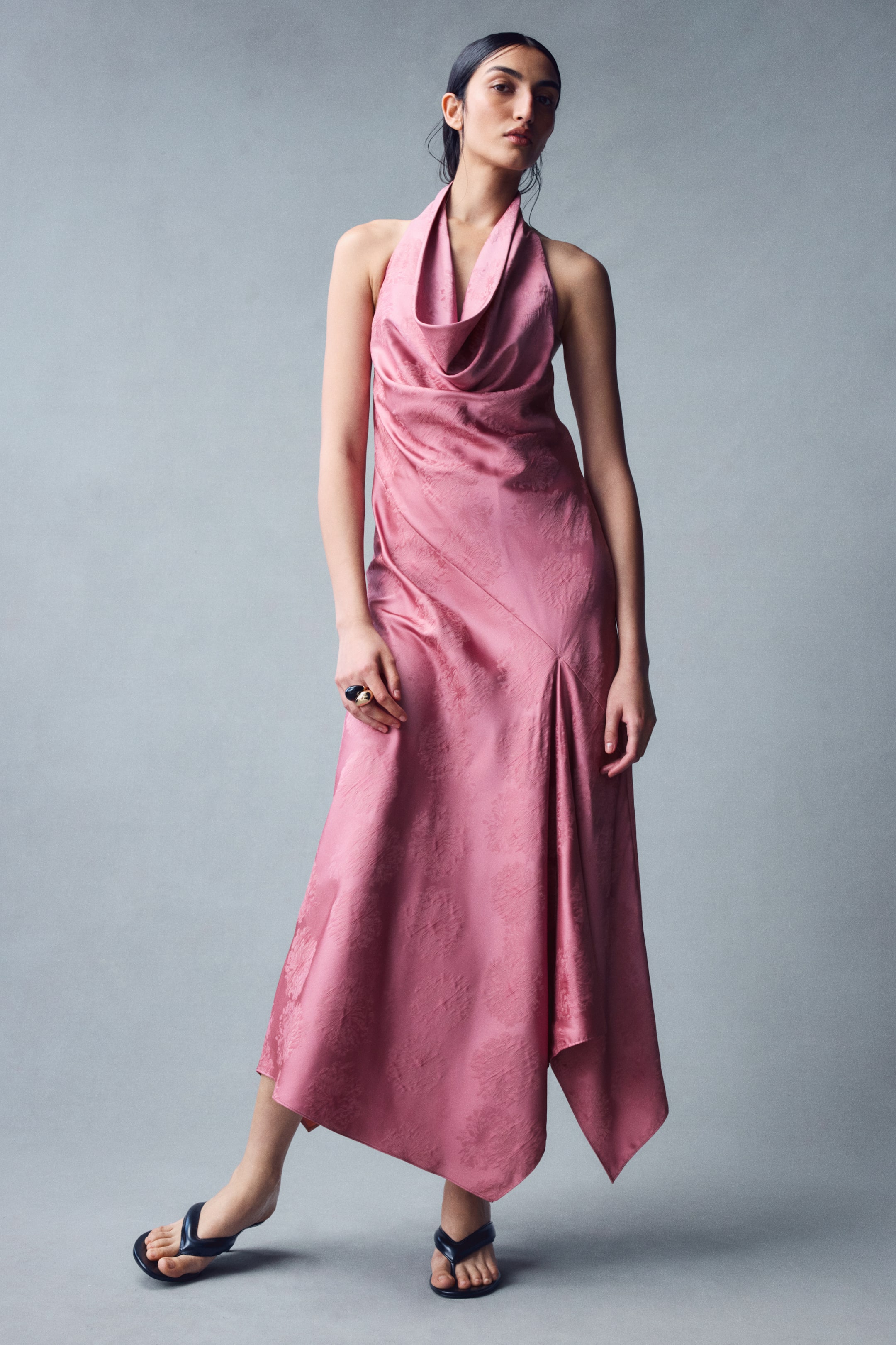 View larger image: The model is wearing an ankle-length pink halterneck dress in a textured, glossy woven fabric. The dress features a deep cowl neckline, a diagonal seam at the front waist, and an asymmetrical skirt with a handkerchief hem.