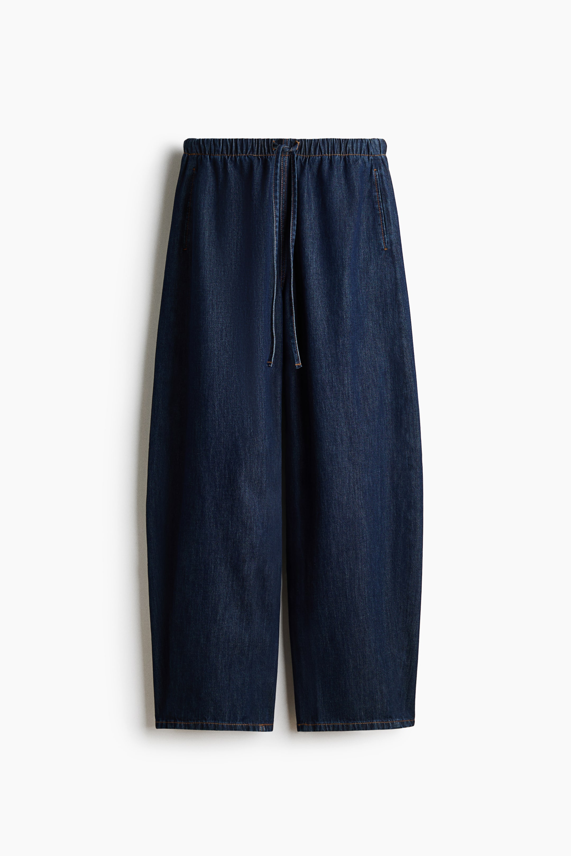 View larger image: Dark blue denim pants are shown front-facing, featuring a relaxed, wide-leg fit with an elasticated waistband and a central drawstring closure. They feature side pockets and contrast orange stitching throughout.
