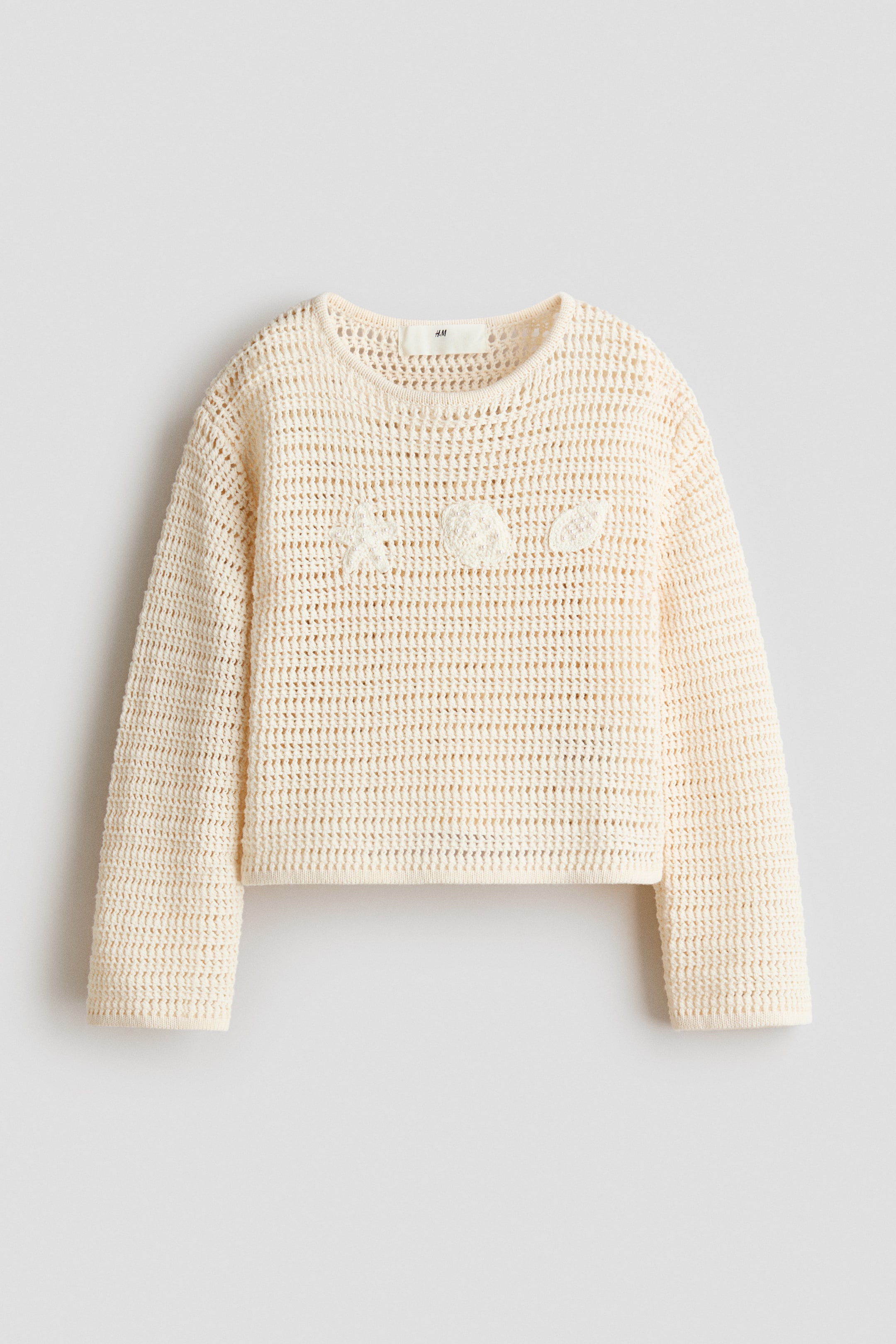 Größeres Bild ansehen: Ein cremefarbener, gehäkelter Pullover wird von vorne gezeigt, mit einem Rundhalsausschnitt, langen Ärmeln und einem leicht verkürzten, geraden Saum. Drei bestickte, maritim inspirierte Formen, darunter ein Seestern, eine Muschel und eine Venusmuschel, zieren den Brustbereich vorne.
