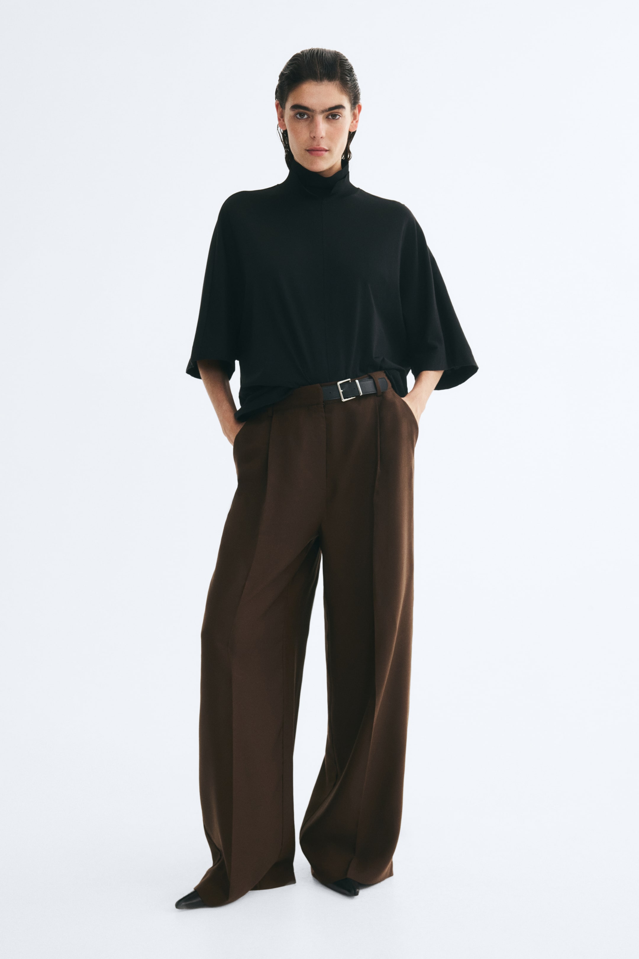 Grotere afbeelding bekijken: De model draagt een bruine wijde broek met persplooien aan de voorkant en staat met de handen in de zijzakken. De broek is gestyled met een ingestopte zwarte top met korte mouwen en opstaande hals en een zwarte riem met zilveren gesp.