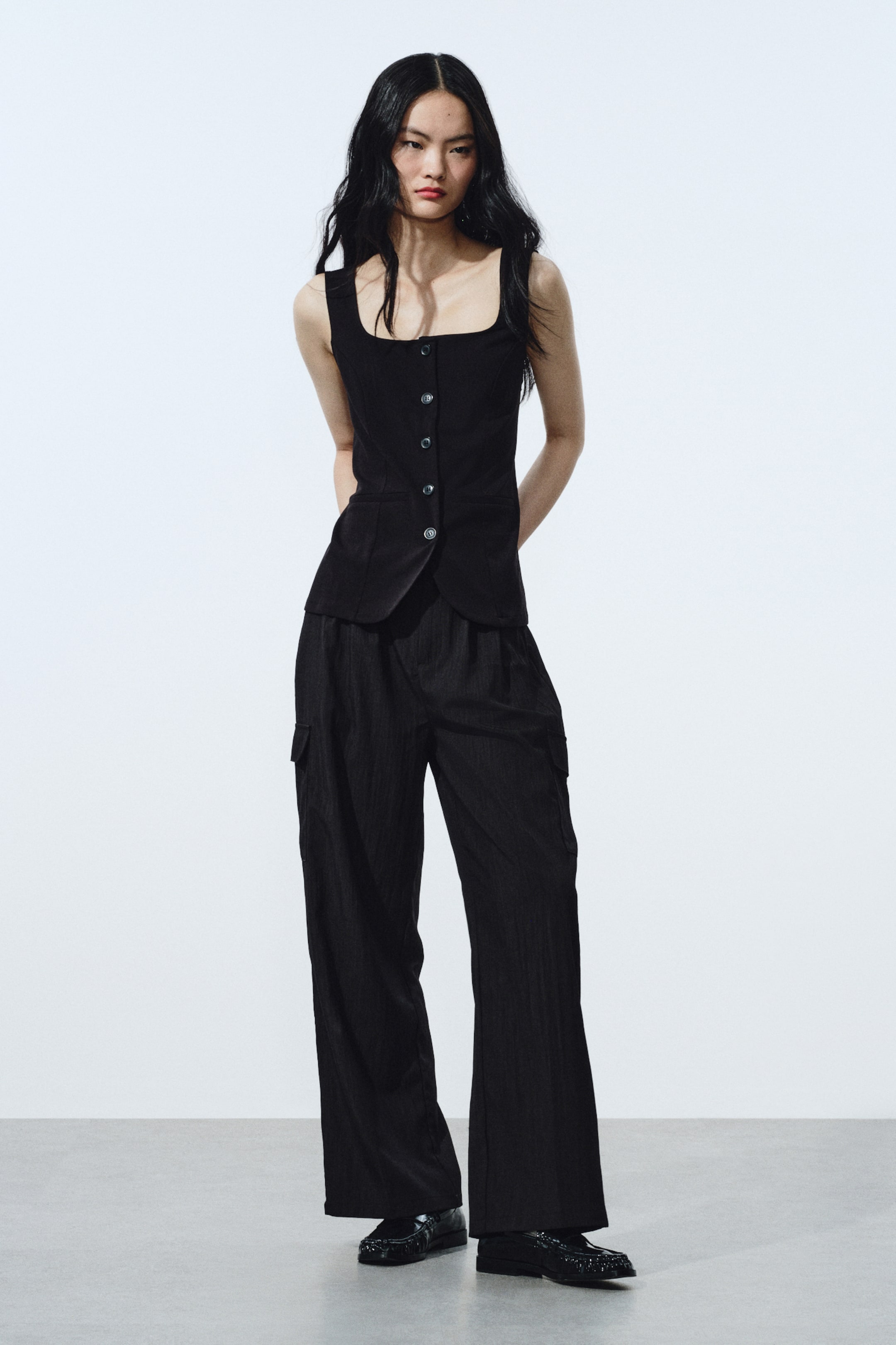 拡大画像を表示: 黒のノースリーブトップはスクエアネック、6つボタンの前開き、2つの小さなフラップポケットを備え、ベスト風の構造を呈しています。黒のワイドレッグカーゴパンツと合わせられており、さりげない縦方向のテクスチャーとサイドのカーゴポケットが特徴です。