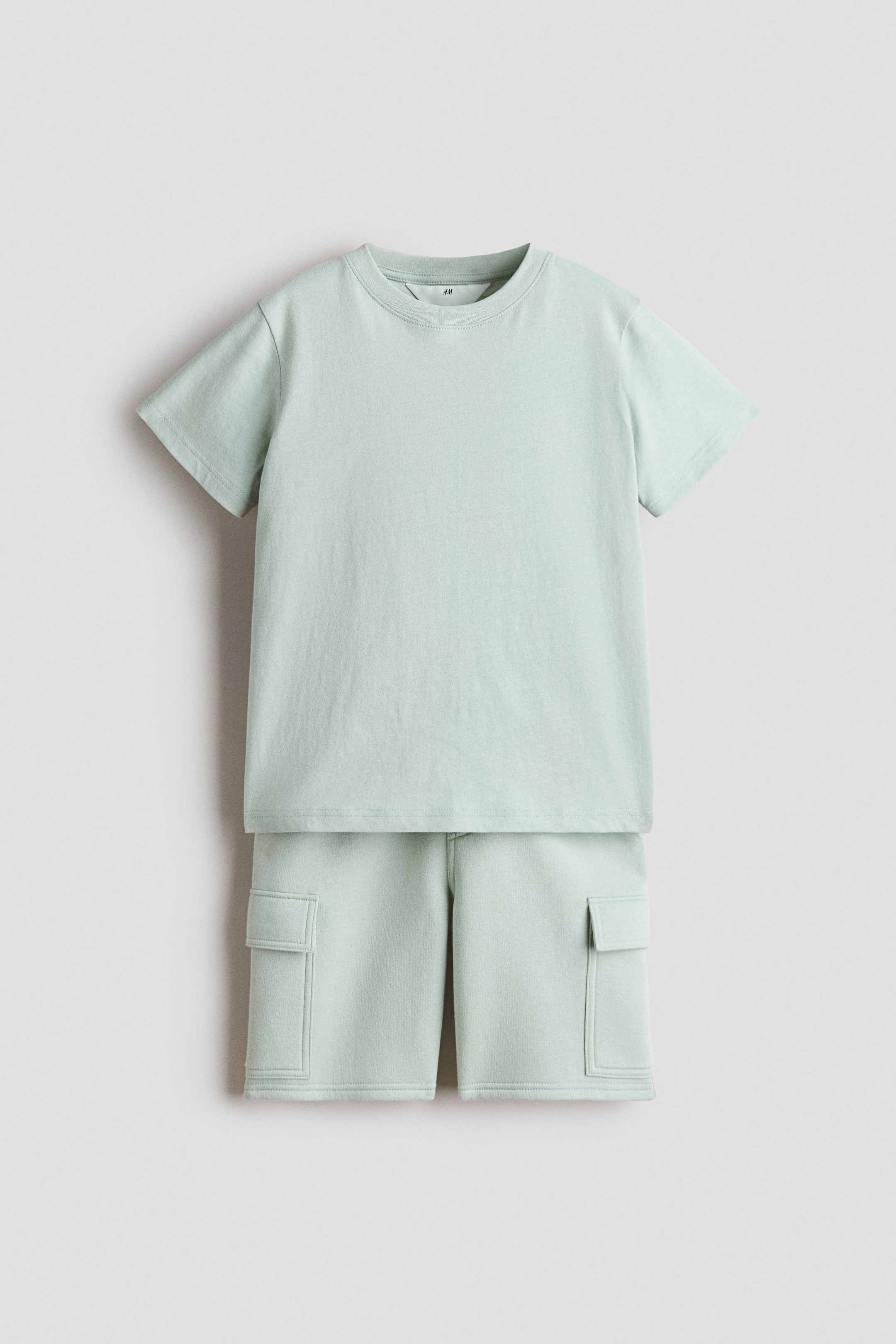 View larger image: A light green T-shirt and shorts set is displayed front-facing, laid flat. The T-shirt features a round, ribbed neckline, short sleeves, and a straight-cut hem, while the shorts boast an elasticated, drawstring waist, a faux fly, diagonal side pockets, and patch leg pockets with flaps.