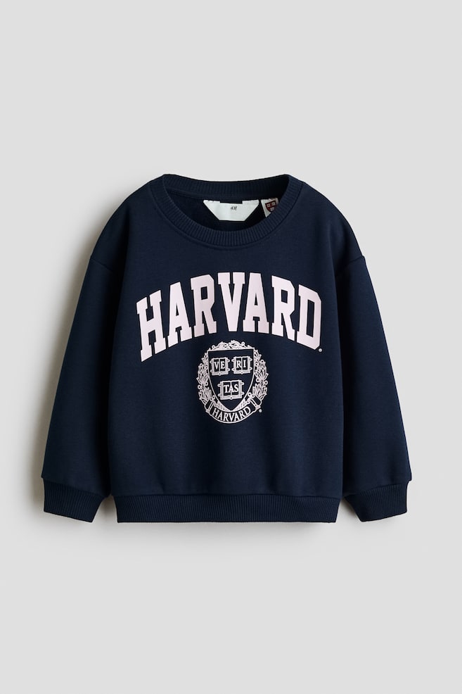 A navy blue sweatshirt is shown from the front, featuring a round ribbed neckline, dropped shoulders, and long sleeves with ribbed cuffs and hem. The chest displays a light pink printed design, with 'HARVARD' arched over a university crest. - 1