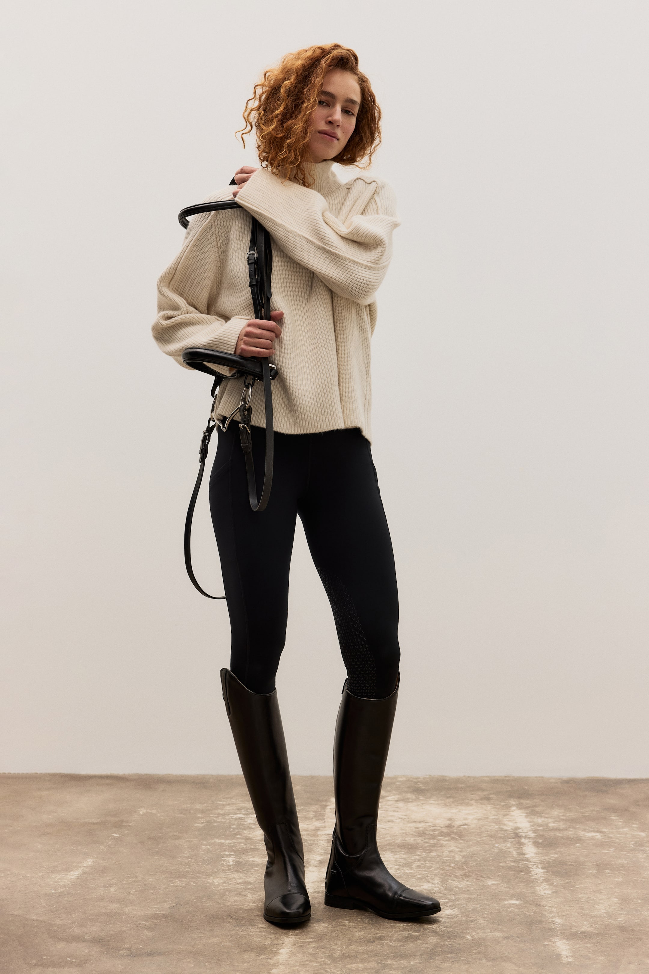 Größeres Bild anzeigen: Das Produkt ist ein cremefarbener, gerippter Strickpullover mit hohem Kragen und weiten Ärmeln, schwarze, figurbetonte Reithosen mit strukturierten Grip-Panels und hohe, polierte schwarze Reitstiefel. Ein schwarzes Lederzaumzeug ruht über einer Schulter.
