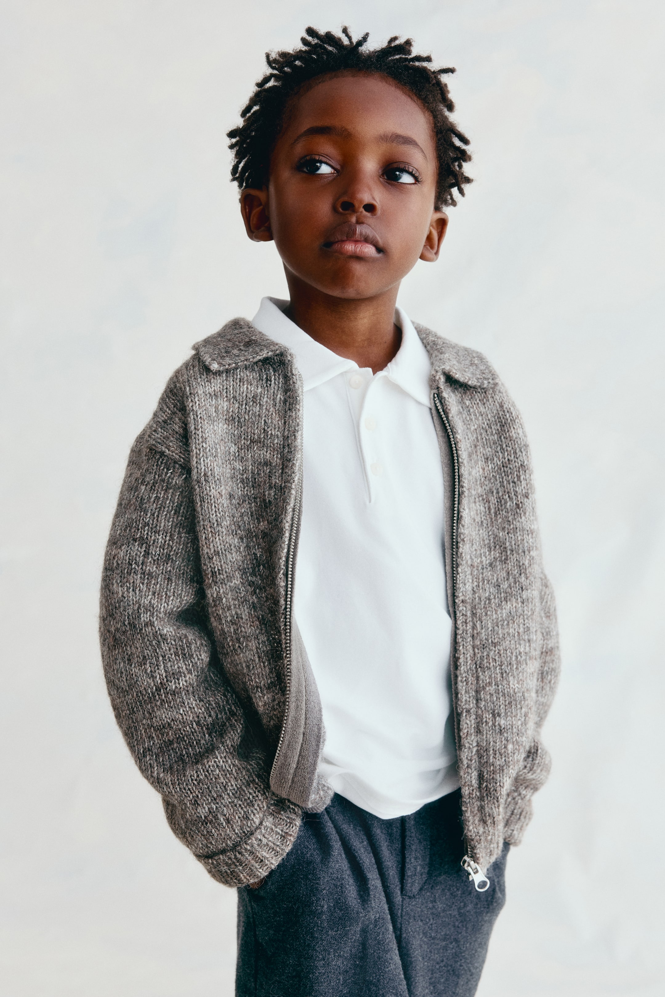 View larger image: A grey-brown mélange knit cardigan is shown from the front, featuring a classic collar, dropped shoulders, and ribbed cuffs. The cardigan is worn unzipped, layered over a white polo shirt to showcase its soft, textured fabric and relaxed fit.