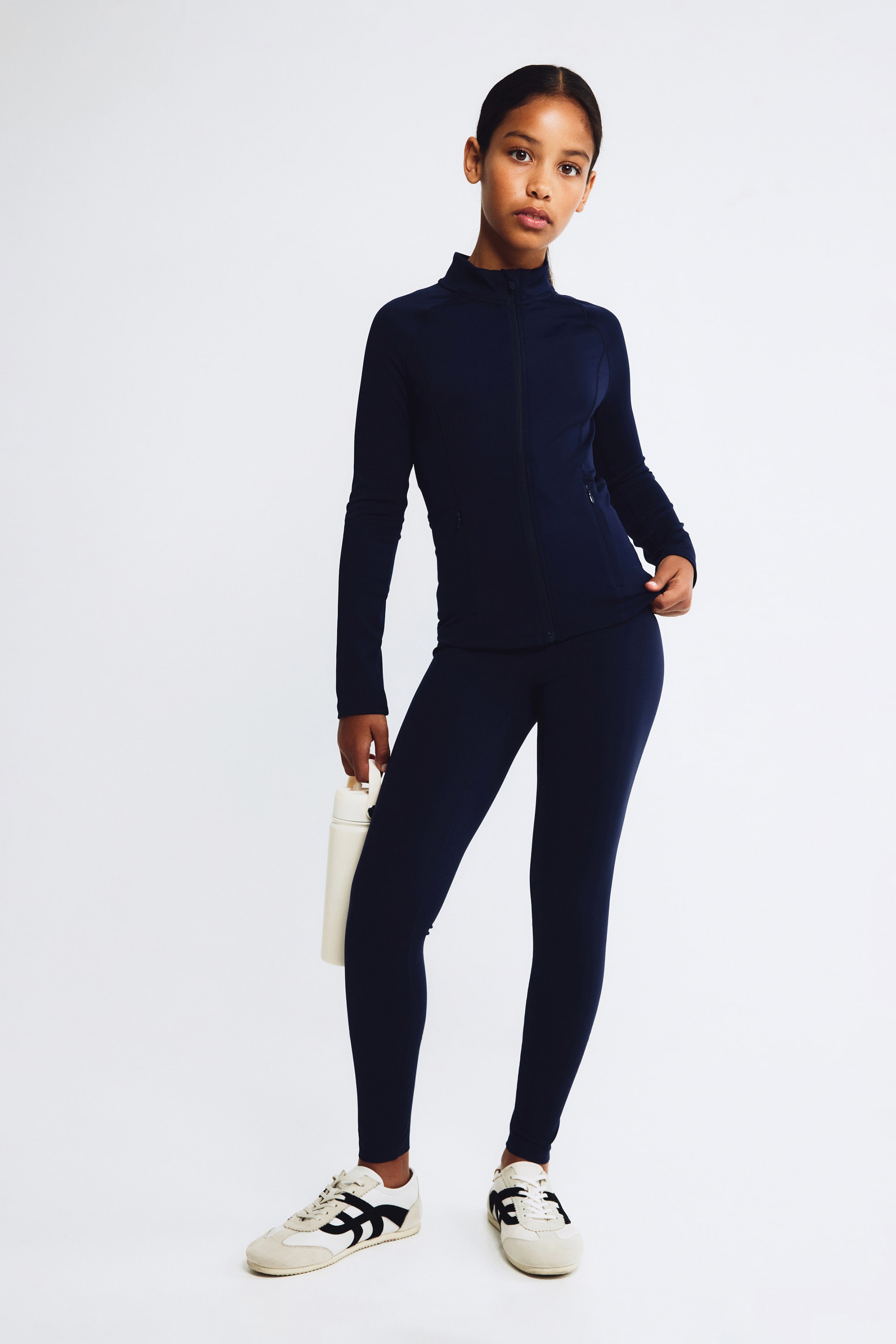 Grösseres Bild ansehen: Ein marineblaues Activewear-Set ist von vorne zu sehen und besteht aus einer taillierten Langarmjacke mit hohem Stehkragen und Frontreissverschluss sowie dazu passenden, körpernahen Leggings in voller Länge.