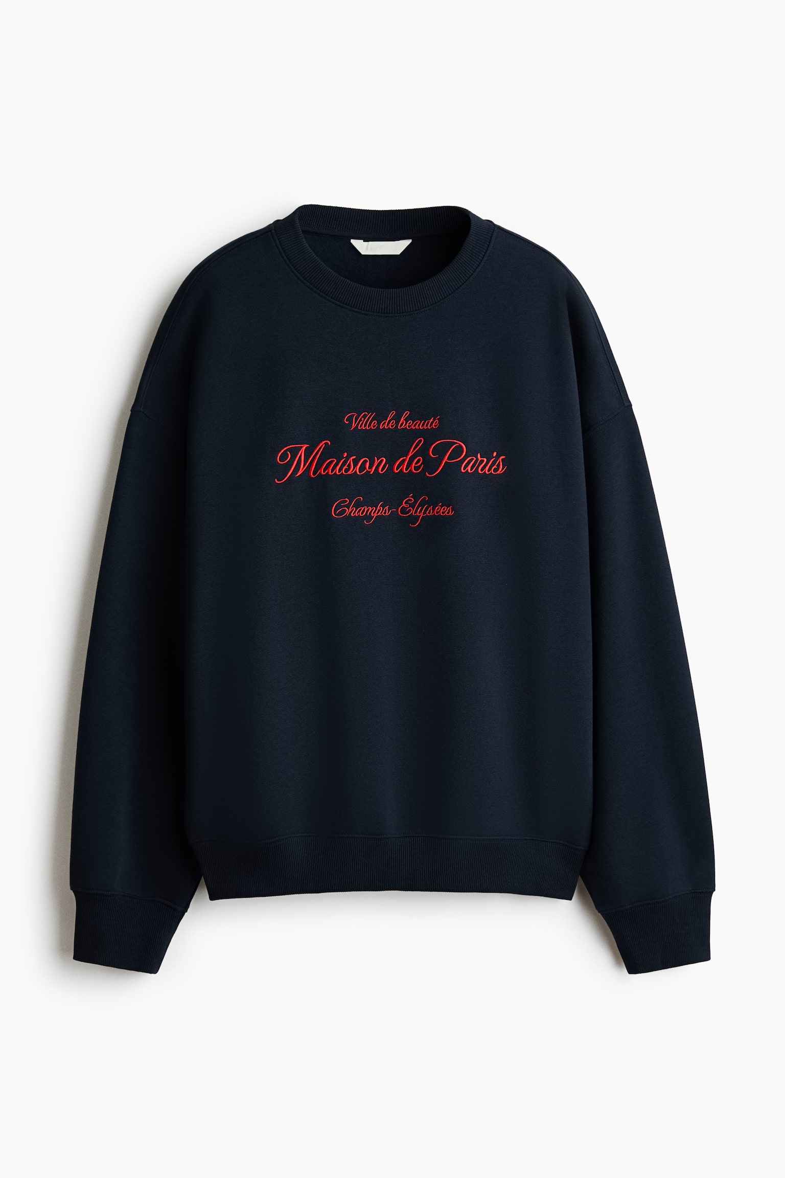 A navy blue loose-fit sweater in brushed fleece fabric is shown from the front, featuring a round, rib-trimmed neckline, dropped shoulders, and ribbed cuffs and hem. The front of the sweater displays red cursive embroidered text reading "Ville de beautÃ©", "Maison de Paris", and "Champs-ÃlysÃ©es".
