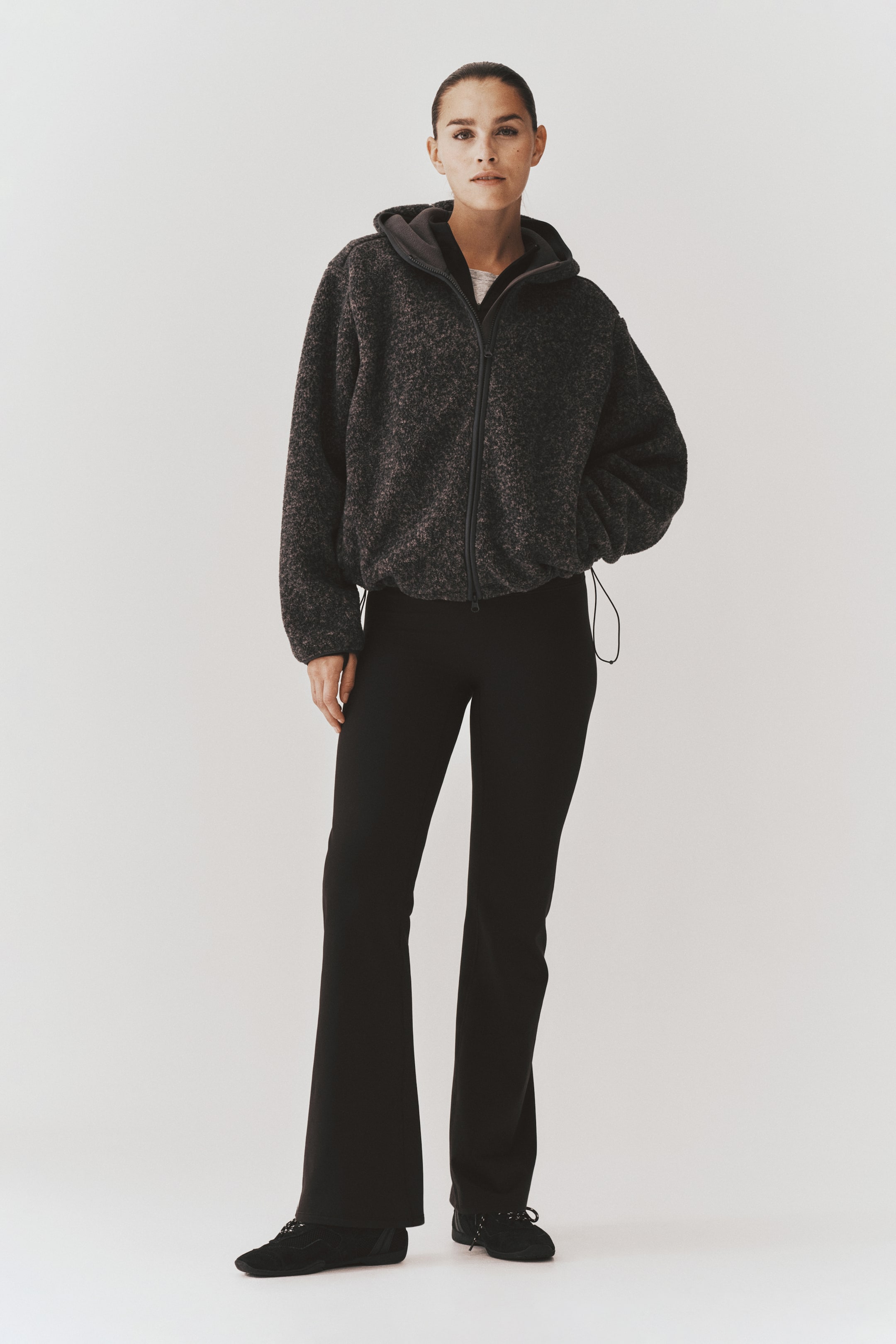 Ver imagen más grande: La modelo lleva una sudadera con capucha de forro polar texturizado en gris oscuro, con cremallera frontal, capucha y bajo con cordón ajustable. La prenda exterior se combina con pantalones negros acampanados y zapatillas negras.