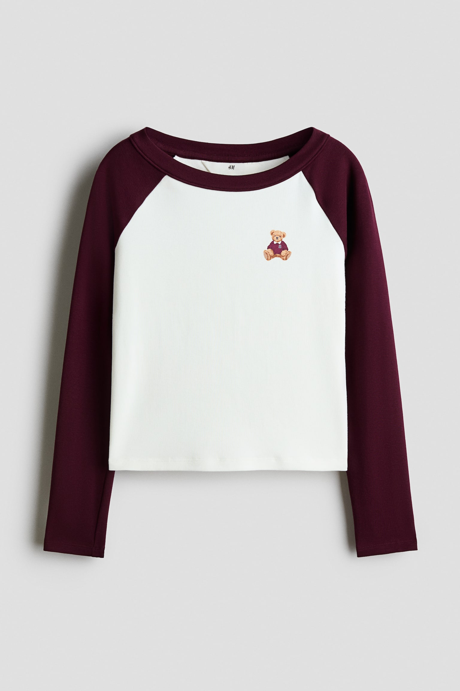 A front-facing, long-sleeve raglan top is featuring a white body with contrasting burgundy sleeves and a matching ribbed crew neck. A small embroidered teddy bear wearing a burgundy sweater is detailing the upper left chest. - 1