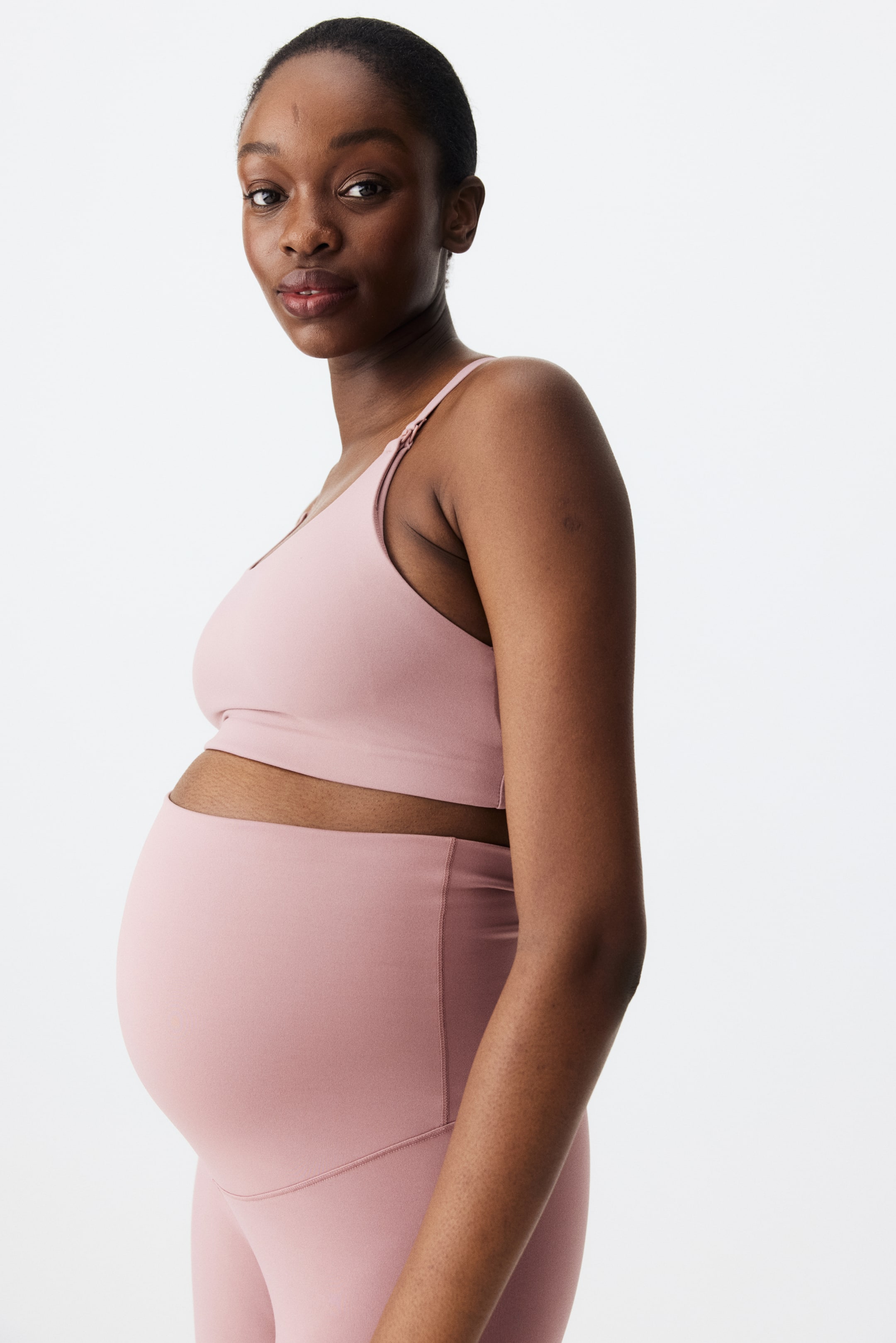 Ampliar la imagen: El conjunto de ropa activa de maternidad rosa presenta un bra deportivo con escote en v, tirantes delgados ajustables y un broche visible para lactancia. Los leggings de talle alto completan el conjunto, diseñados para acomodar un vientre de embarazada, con ambas piezas ajustándose cómodamente y mostradas desde una vista lateral ligeramente angular.