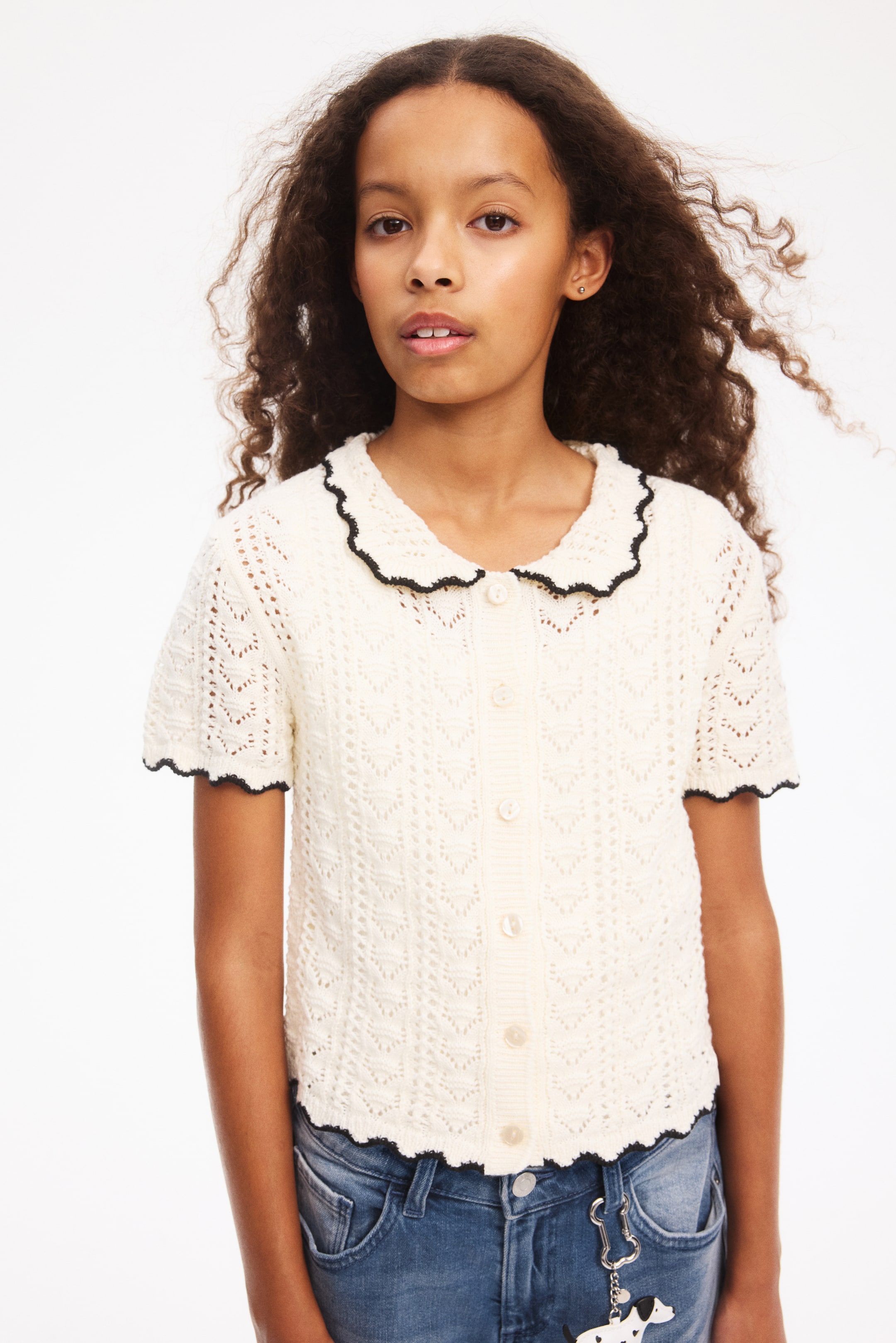 View larger image: A short-sleeved, cream-coloured cardigan features a hole-knit pattern with a crocheted look and is shown from the front. It has a collar with black scalloped, ribbed edges, buttons down the front with a ribbed button band, and black scalloped, ribbed edges on the cuffs and hem.
