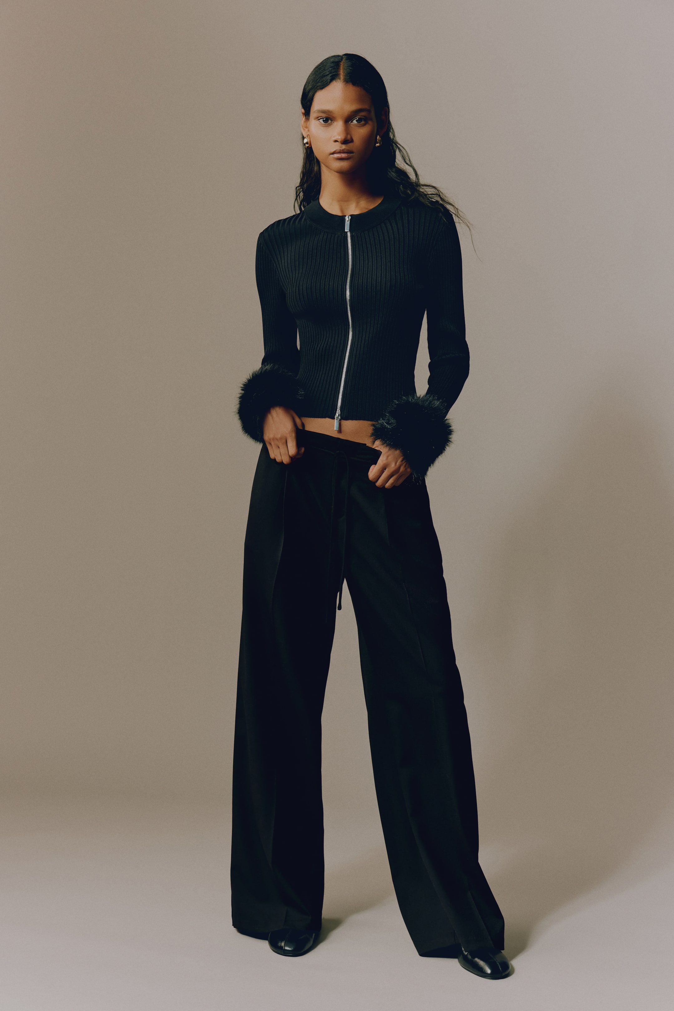 View larger image: A black ribbed knit zip-up top featuring a silver-tone front zip and long sleeves with faux fur cuffs, paired with black wide-leg trousers showcasing a drawstring waist.