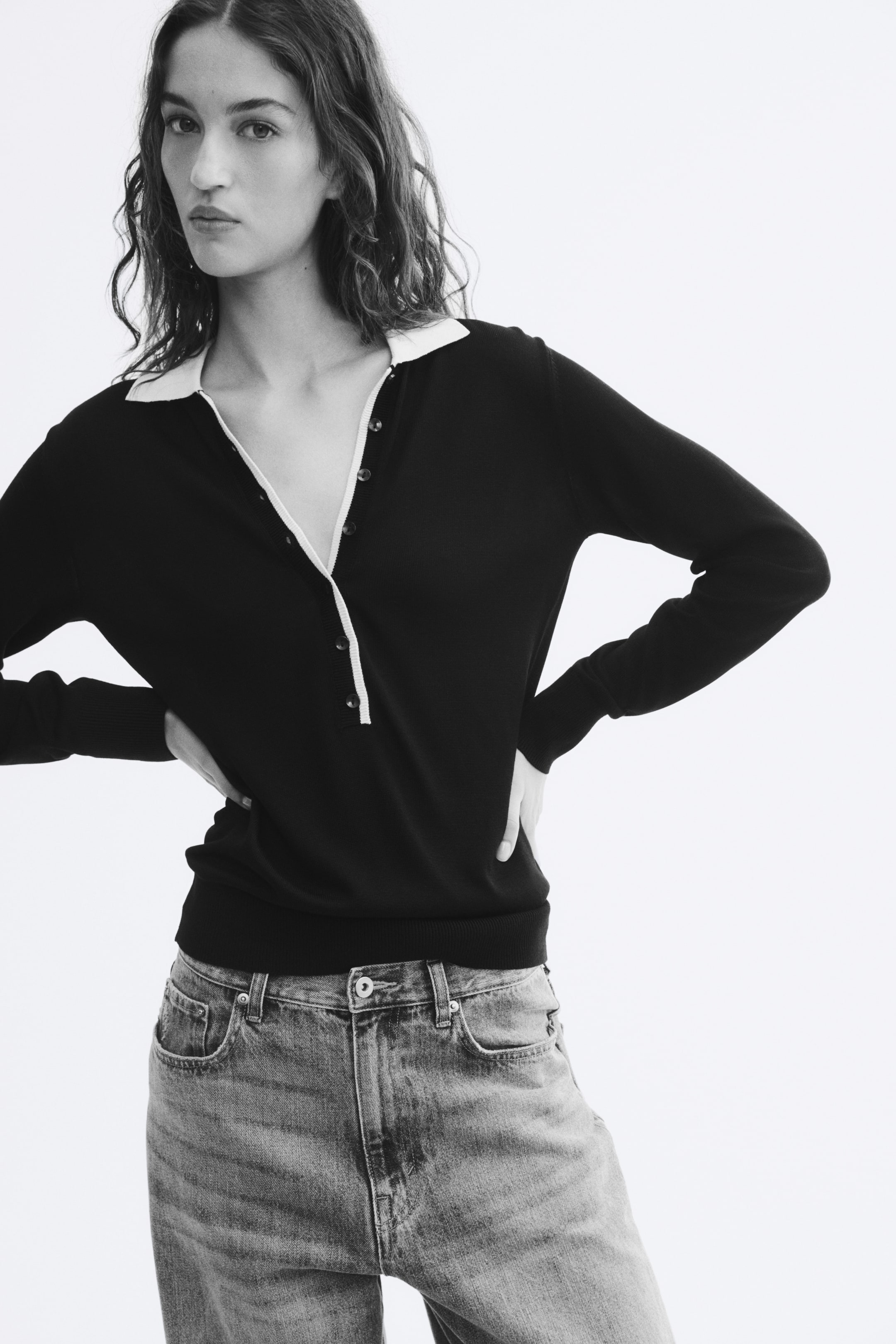 View larger image: A fine-knit sweater featuring a contrasting collar, an elongated button placket, and ribbed cuffs is worn by the model. The sweater is styled with denim jeans, with the model posing with hands on hips.