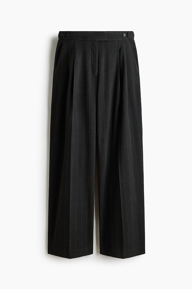 Wide-leg trousers in a dark grey herringbone weave are shown facing front, featuring a high waist with adjustable button tabs at each side and front pleats. Sharp vertical creases are running down the center of each wide leg. - 1
