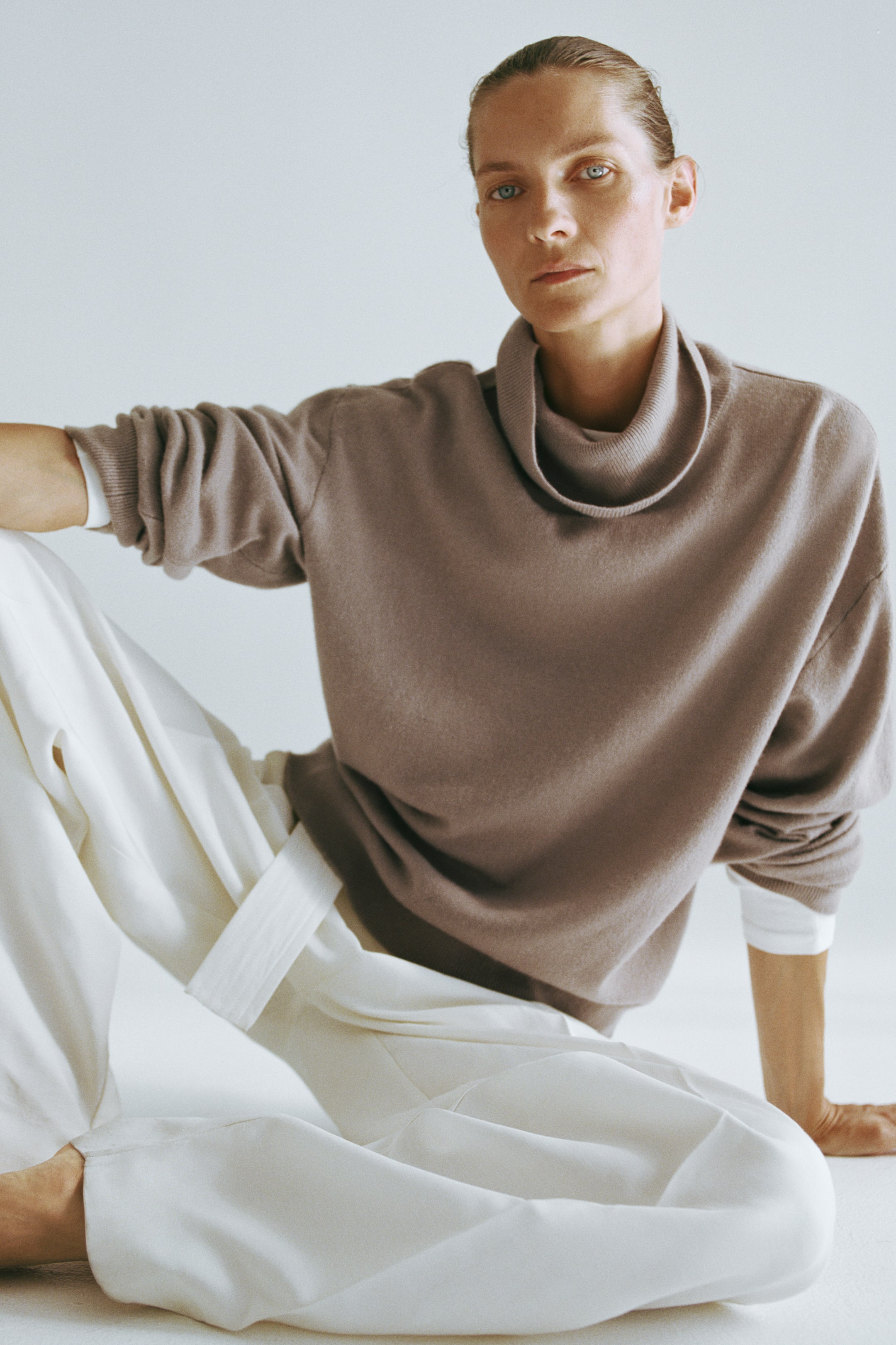 Agrandir l'image: Un pull oversize en cachemire taupe, doté d’un col roulé drapé et d’épaules tombantes, est porté par le mannequin. Les manches longues sont retroussées pour dévoiler une sous-couche blanche, et le pull est associé à un pantalon blanc à jambes larges.