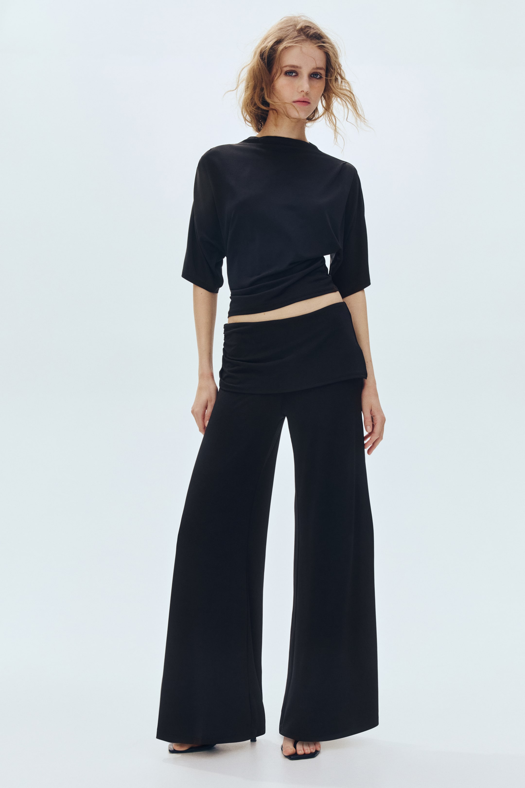 View larger image: A black, loose-fitting top with elbow-length sleeves, a high neckline, and a ruched hem is styled to reveal the midriff, paired with black, flowing wide-leg trousers, all visible from a front-facing angle.